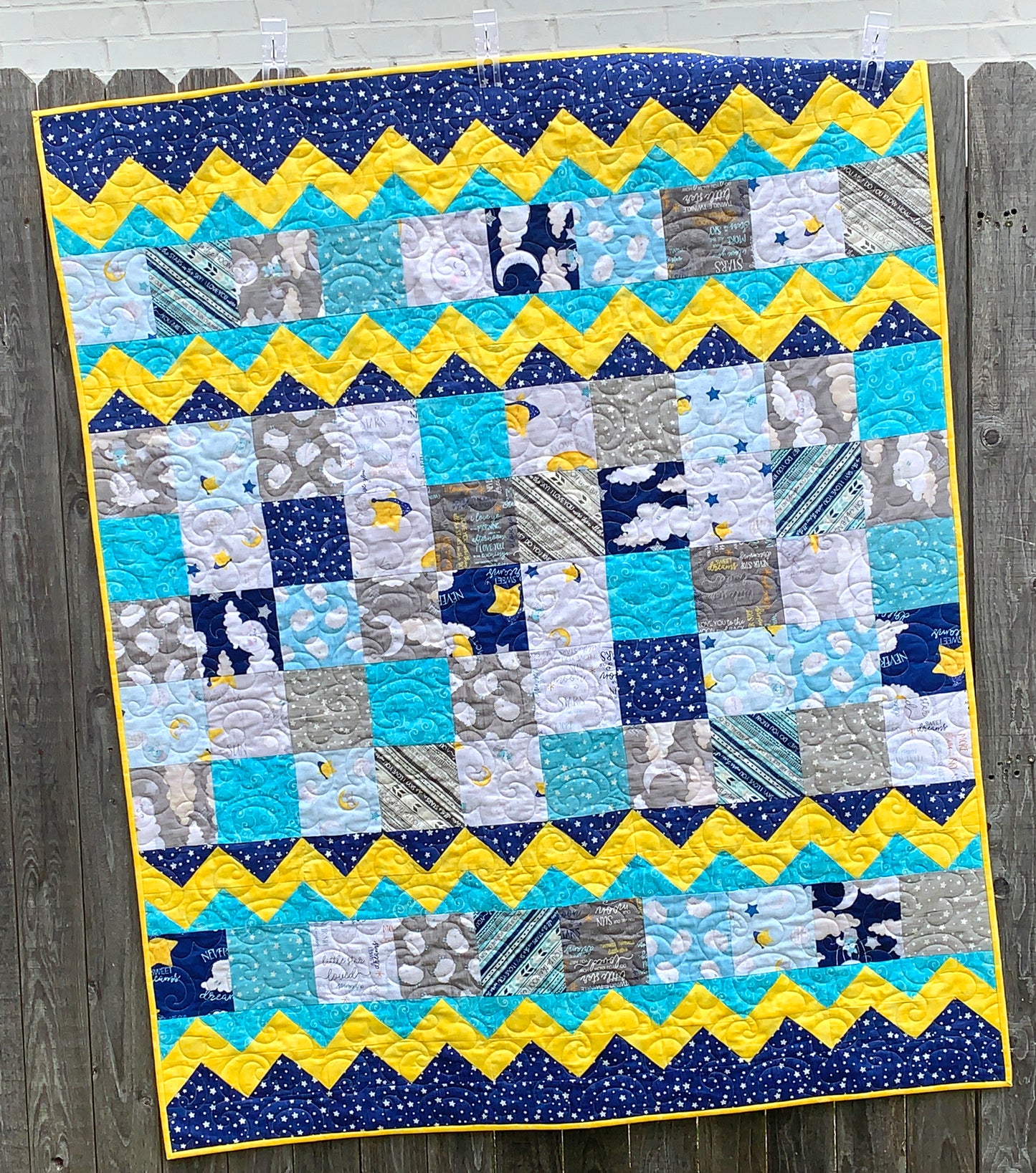 Blue, yellow, teal and gray patchwork baby or toddler quilt with four zig-zag rows accenting the patchwork blocks. Quilt is shown displayed on a fence.
