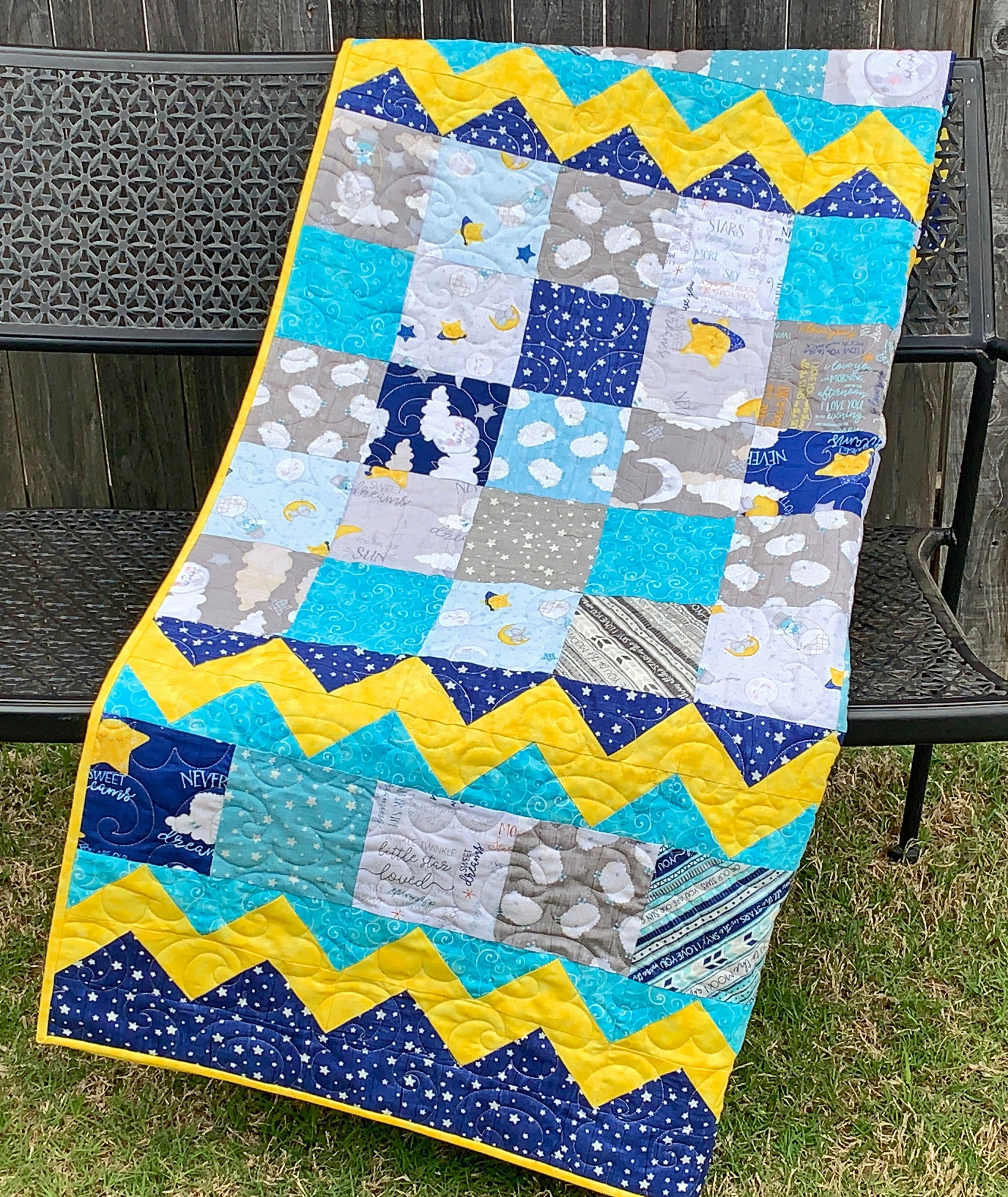Baby Blocks charm square baby quilt pattern with yellow top and bottom zig-zag rows that look like rick-rack with rows of patchwork squares in between. Quilt is trimmed in blue and yellow and is shown displayed on a bench.