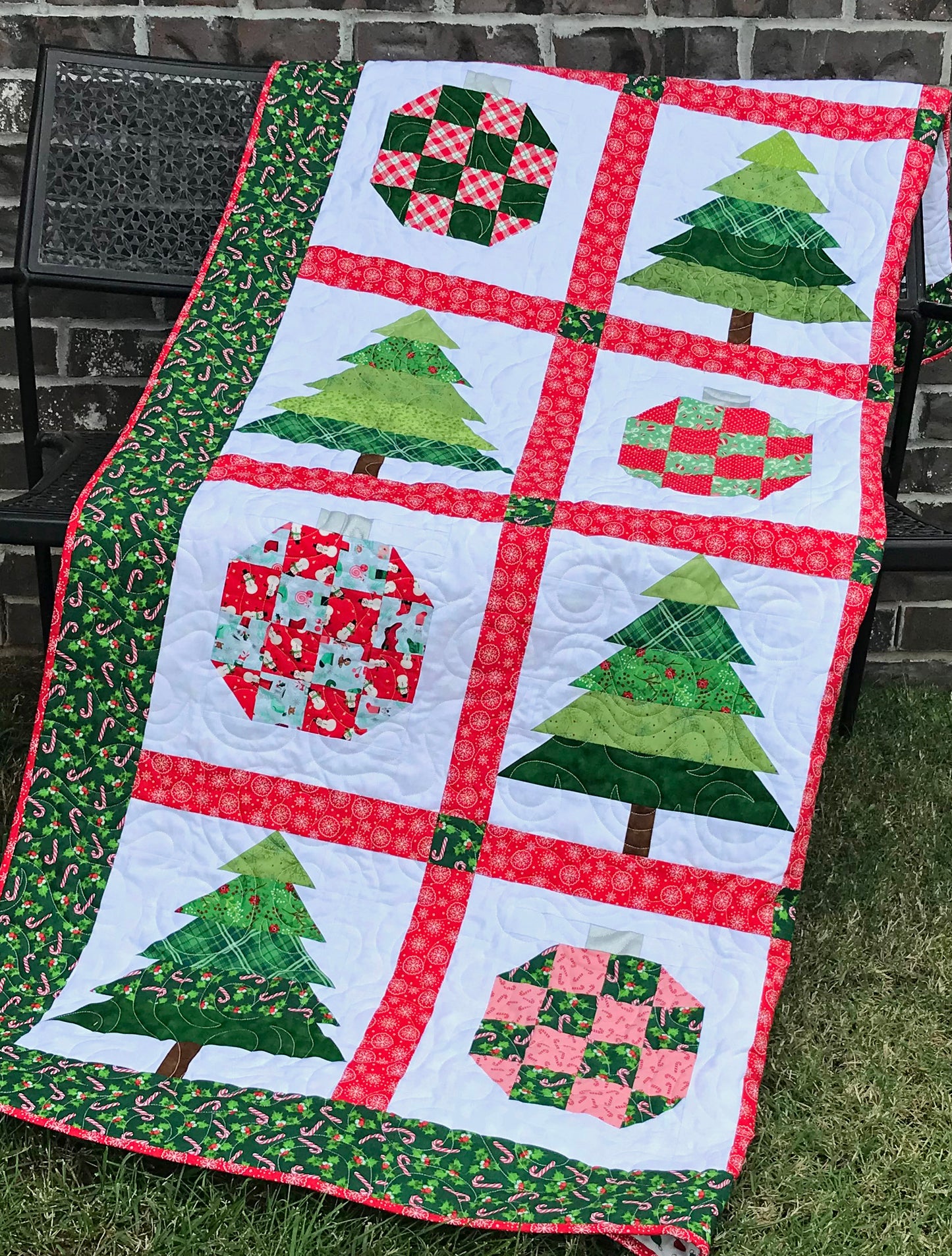 Christmastime quilt pattern with Christmas trees and ornament blocks surrounded by red sashing and a green holly border. Quilt is shown draped across a bench.