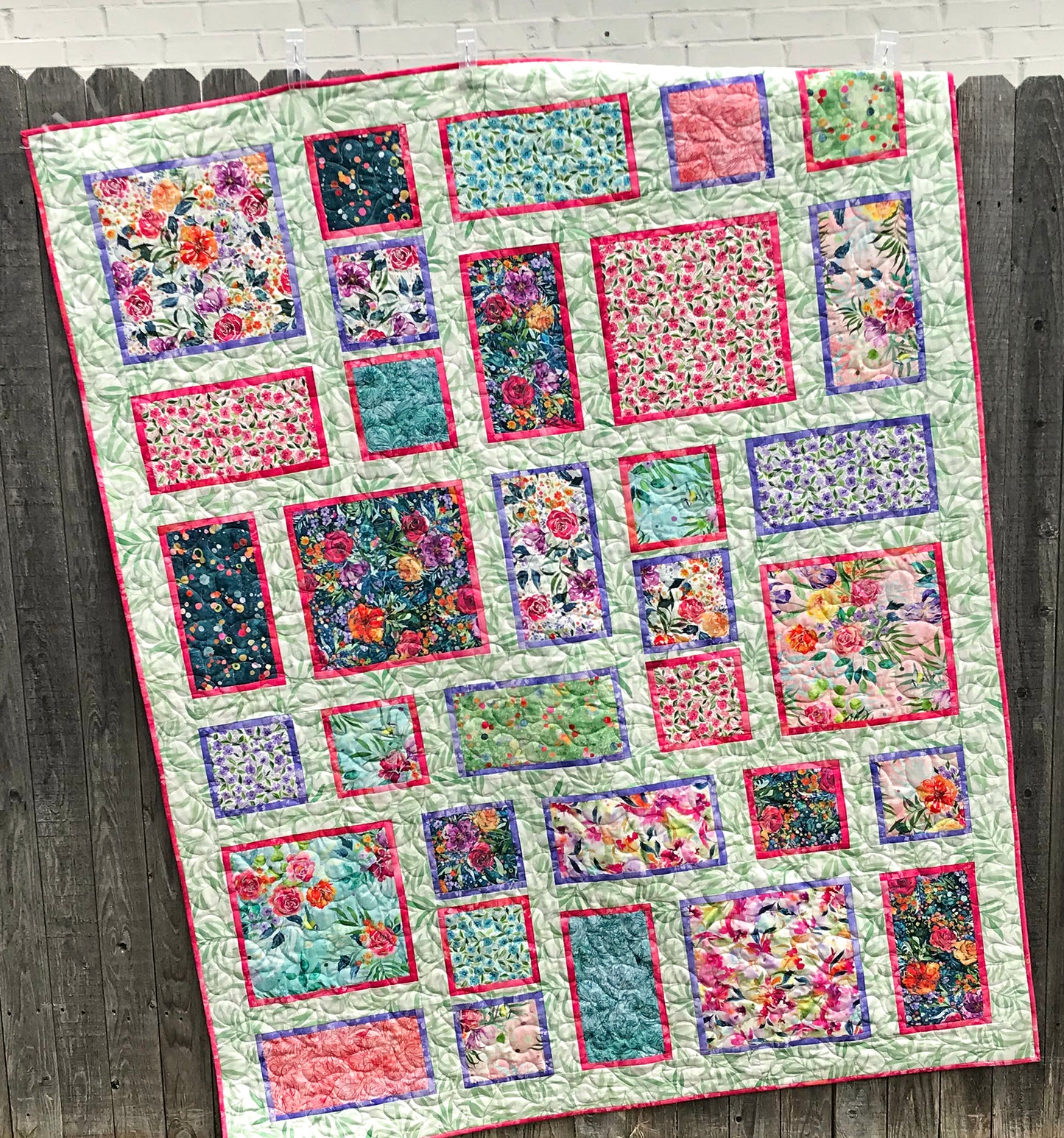 Fancy Frames Quilt pattern of watercolor floral squares and rectangles framed with pink and purple fabric on a light green background.  Quilt shown hanging on a fence.