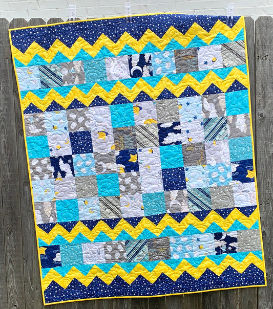 Baby Blocks charm square baby quilt pattern with yellow top and bottom zig-zag rows that look like rick-rack with rows of patchwork squares in between. Quilt is trimmed in blue and yellow and is shown displayed on a fence.