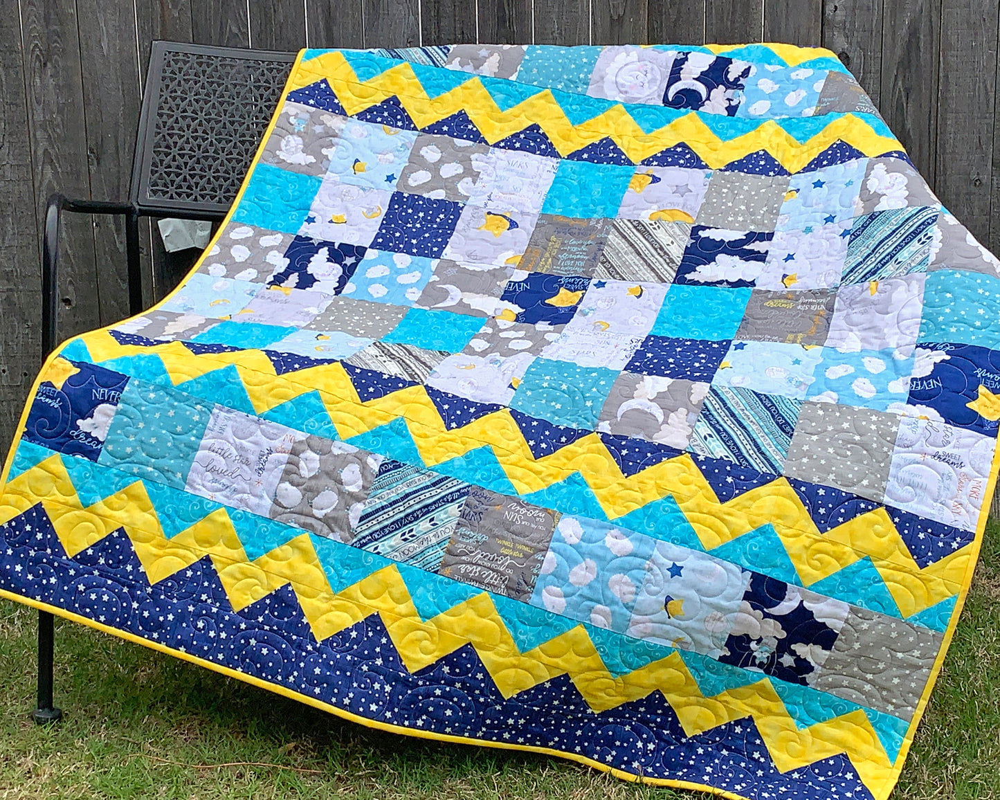 Blue, yellow, teal and gray patchwork baby or toddler quilt with four zig-zag rows accenting the patchwork blocks. Quilt is shown displayed on a bench.