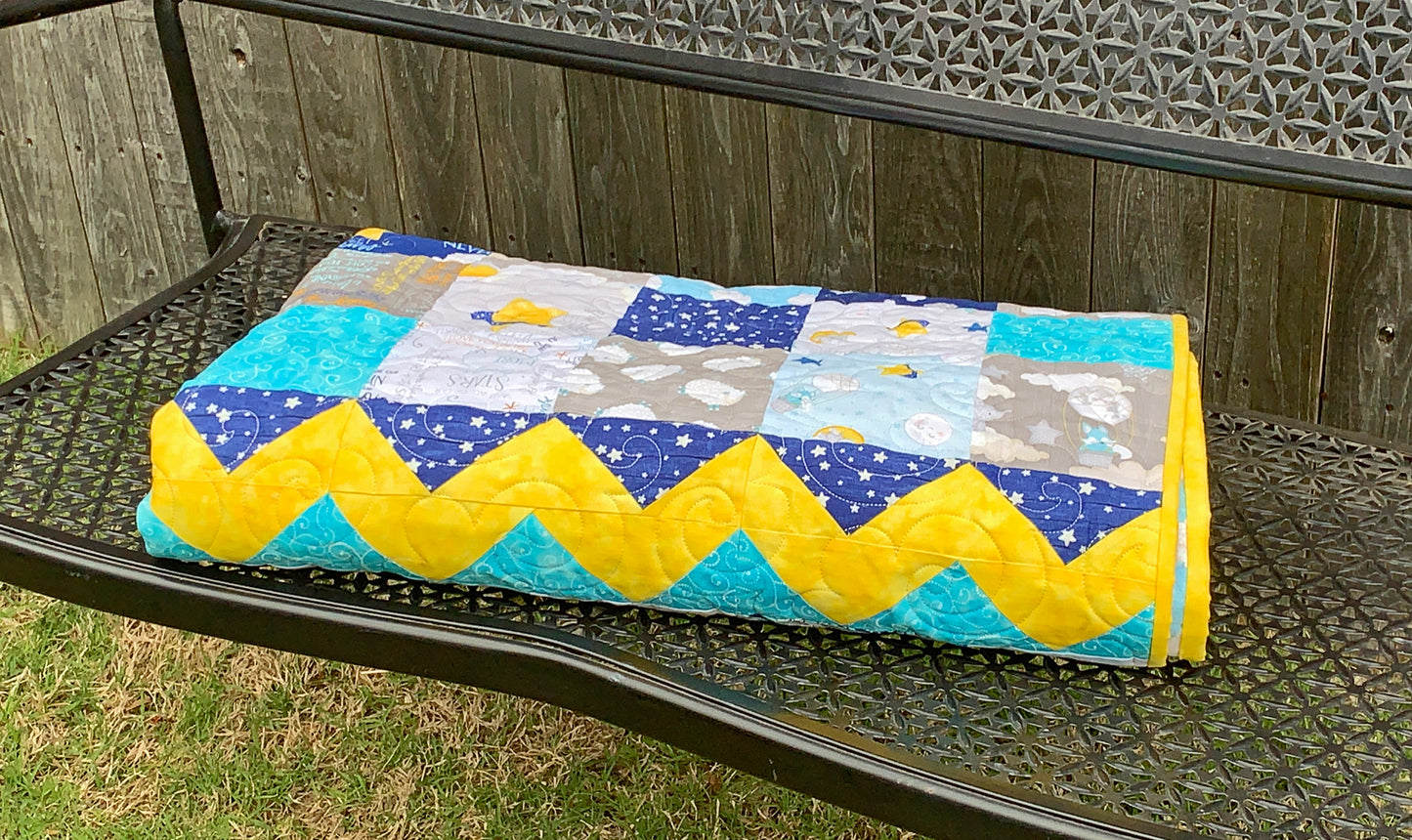 Blue, yellow, teal and gray patchwork baby or toddler quilt with four zig-zag rows accenting the patchwork blocks. Quilt is shown folded on a bench.