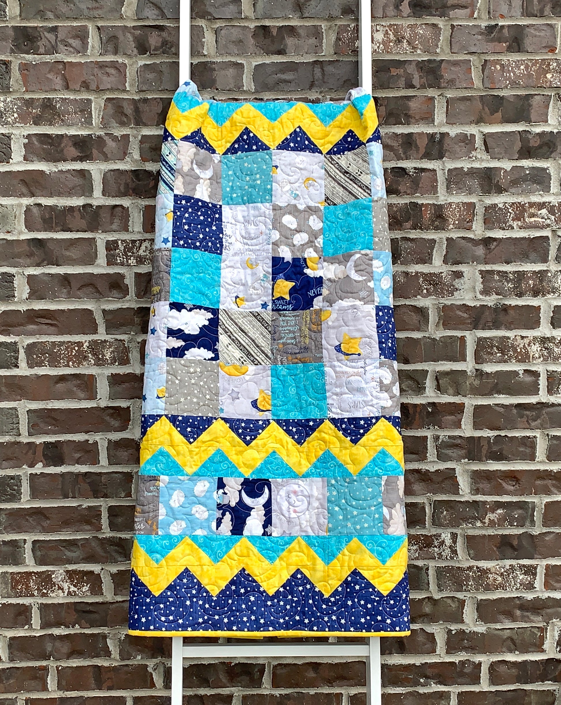 Baby Blocks charm square baby quilt pattern with yellow top and bottom zig-zag rows that look like rick-rack with rows of patchwork squares in between. Quilt is trimmed in blue and yellow and is shown displayed on a ladder.