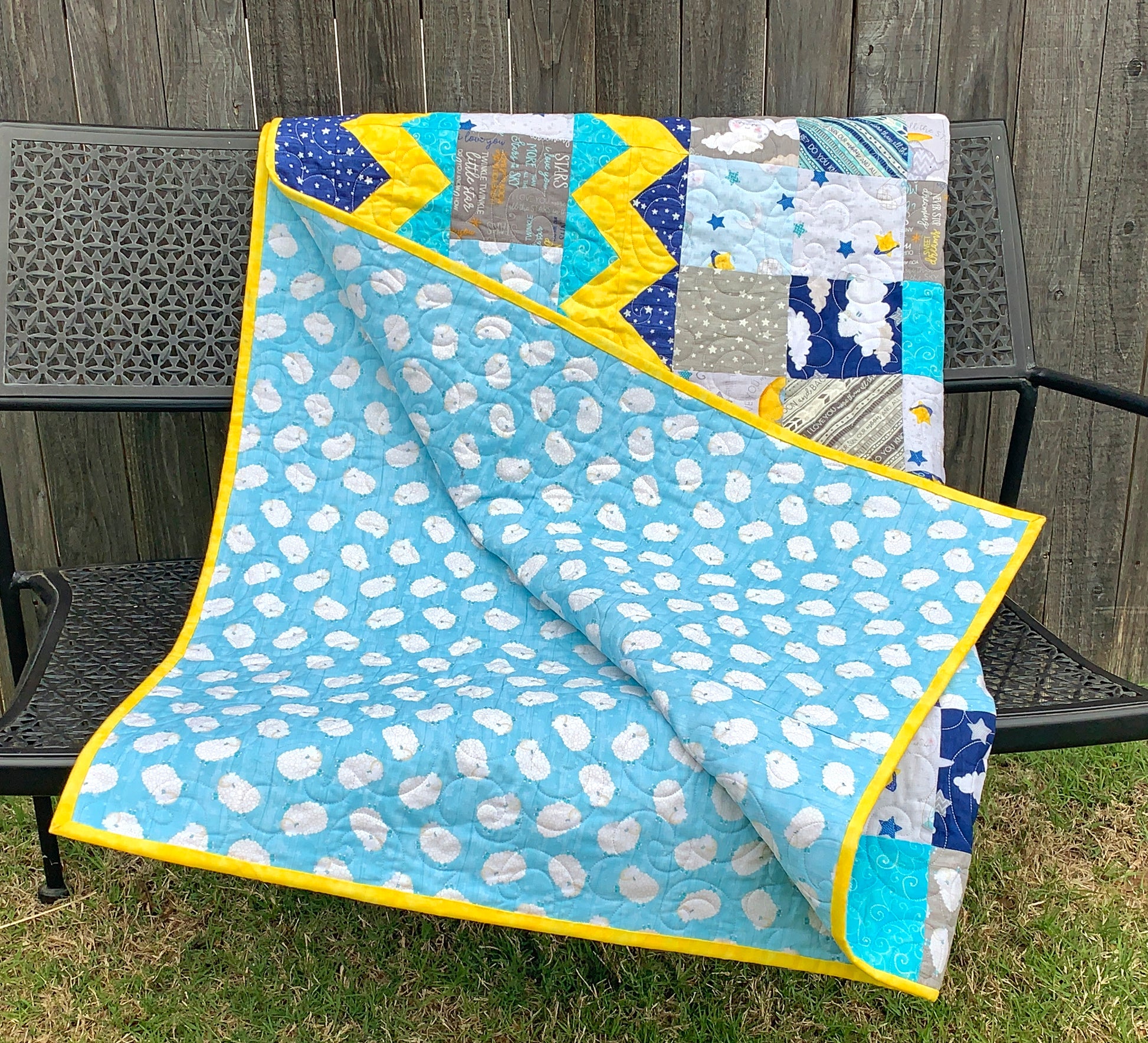 Blue, yellow, teal and gray patchwork baby or toddler quilt with four zig-zag rows accenting the patchwork blocks. Quilt is shown folded up with teal and white backing fabric showing.