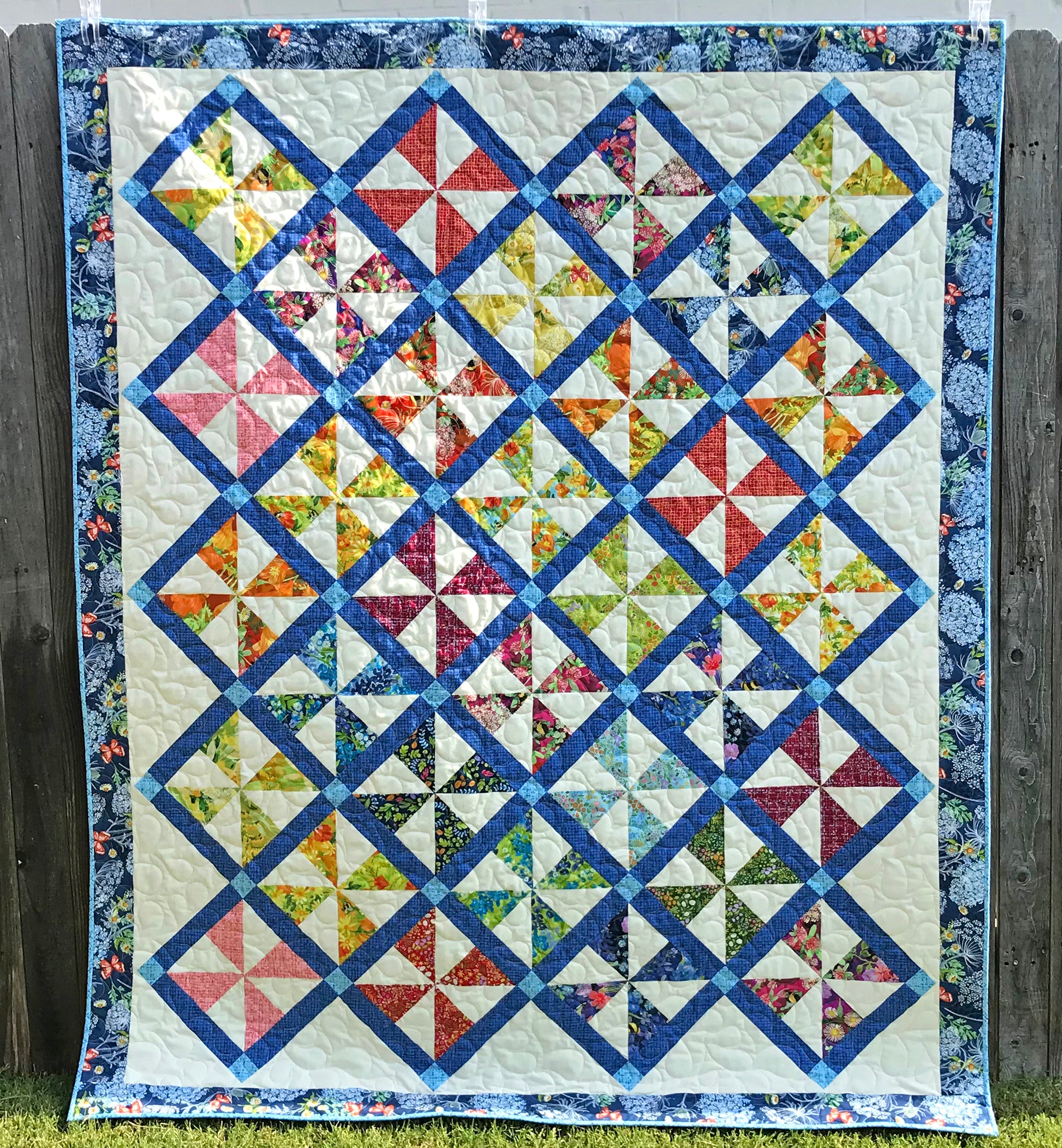 Pinwheel Parade quilt pattern featuring pinwheel blocks set on-point with sashing between the blocks and a floral border. Quilt is shown hanging on a fence.
