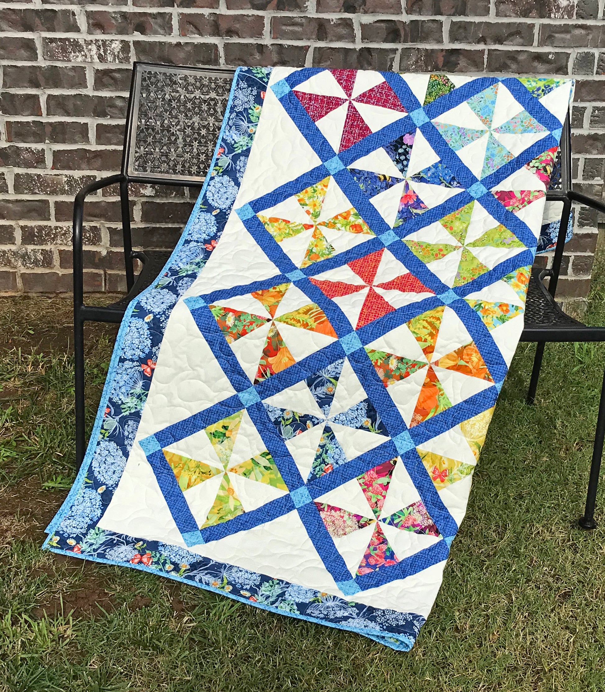 Pinwheel Parade quilt pattern featuring pinwheel blocks set on-point with sashing between the blocks and a floral border. Quilt is shown folded on a bench.