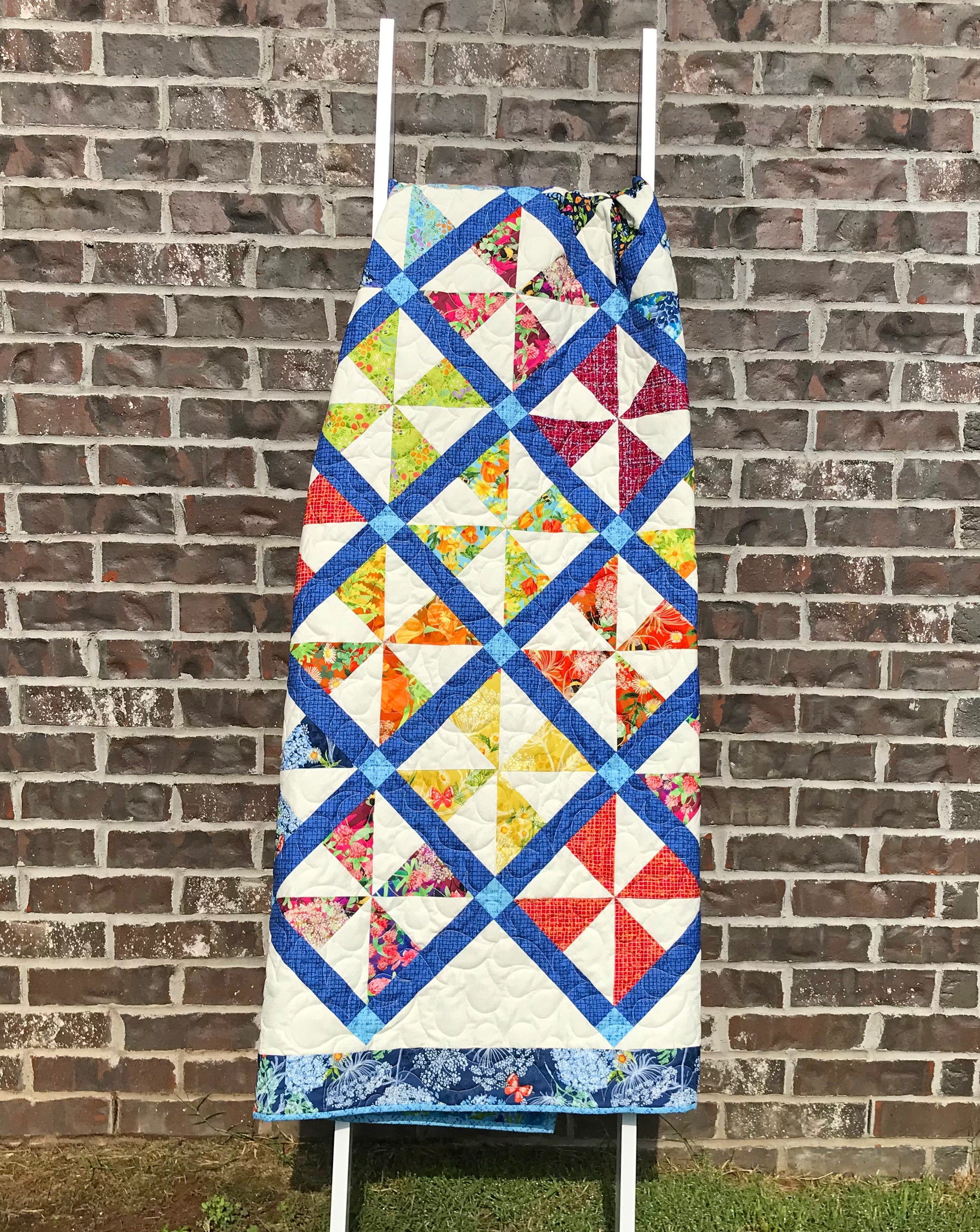 Pinwheel Parade quilt pattern featuring pinwheel blocks set on-point with sashing between the blocks and a floral border. Quilt is shown hanging on a ladder.
