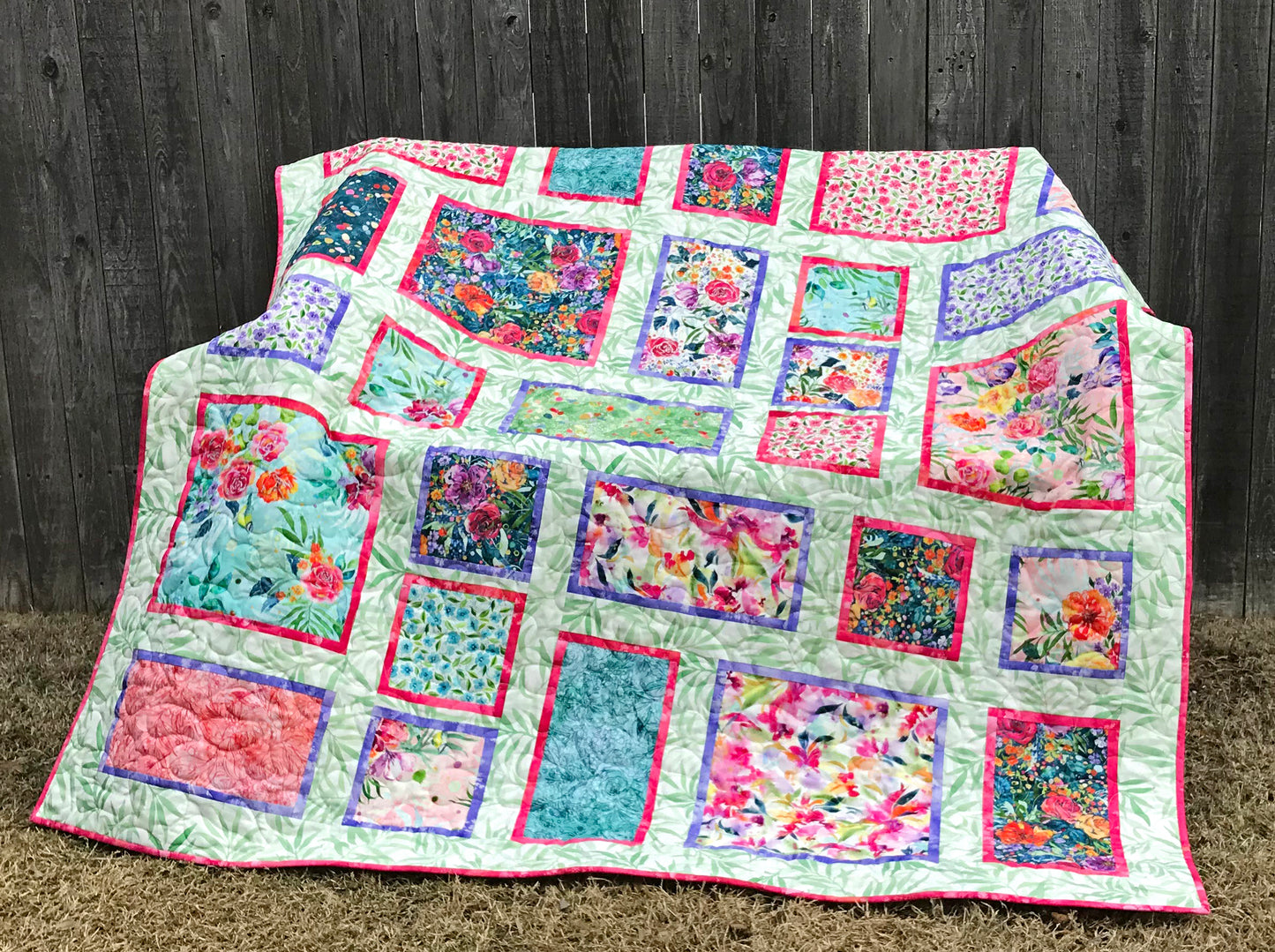 Fancy Frames Quilt pattern of watercolor floral squares and rectangles framed with pink and purple fabric on a light green background. Quilt is shown folded on a bench.