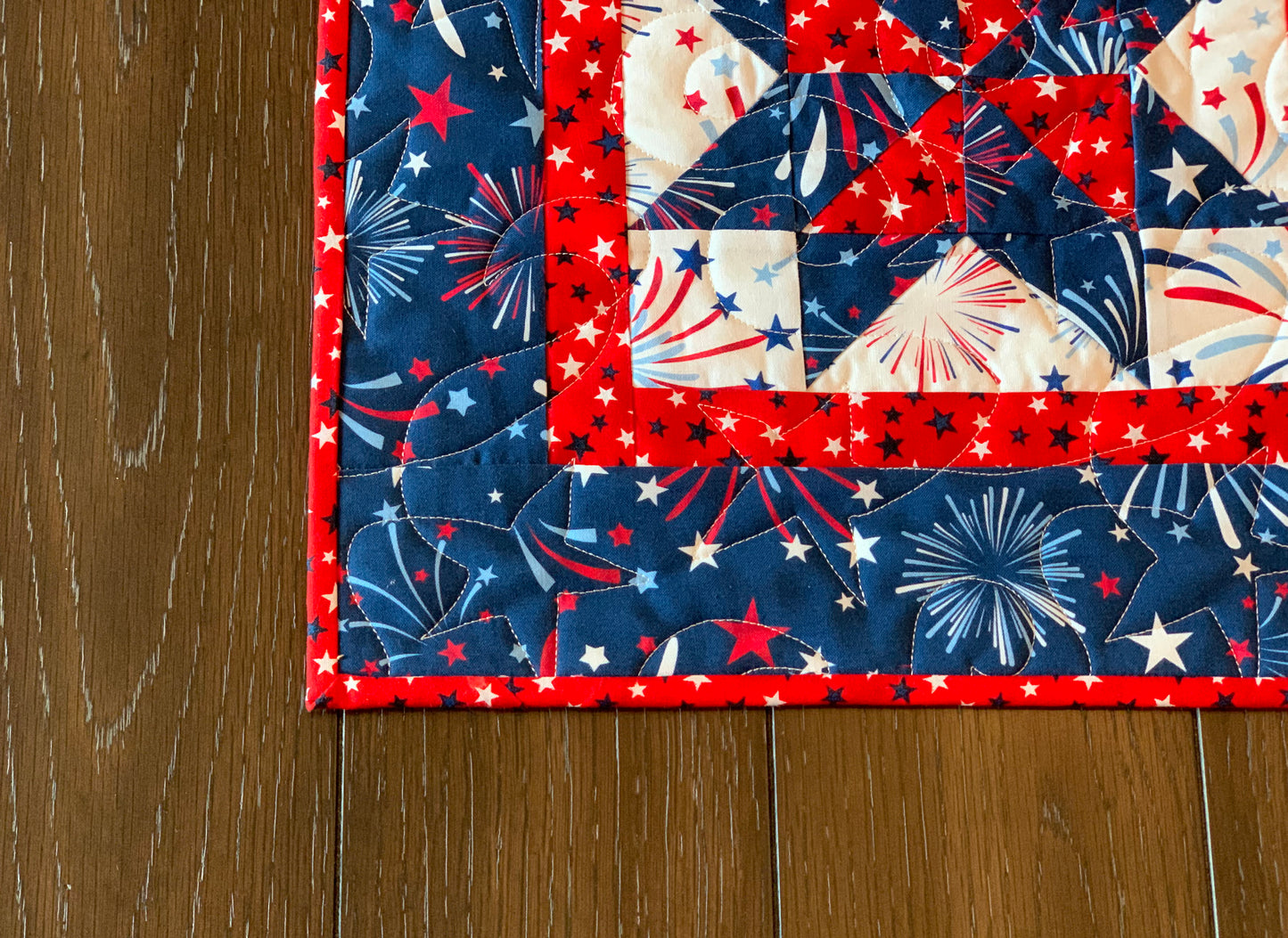 Close up of corner on red white and blue patriotic table runner with four center star blocks that have a pinwheel in the center. The runner has red star sashing between the blocks and a blue fireworks border. Runner is shown displayed on a table.