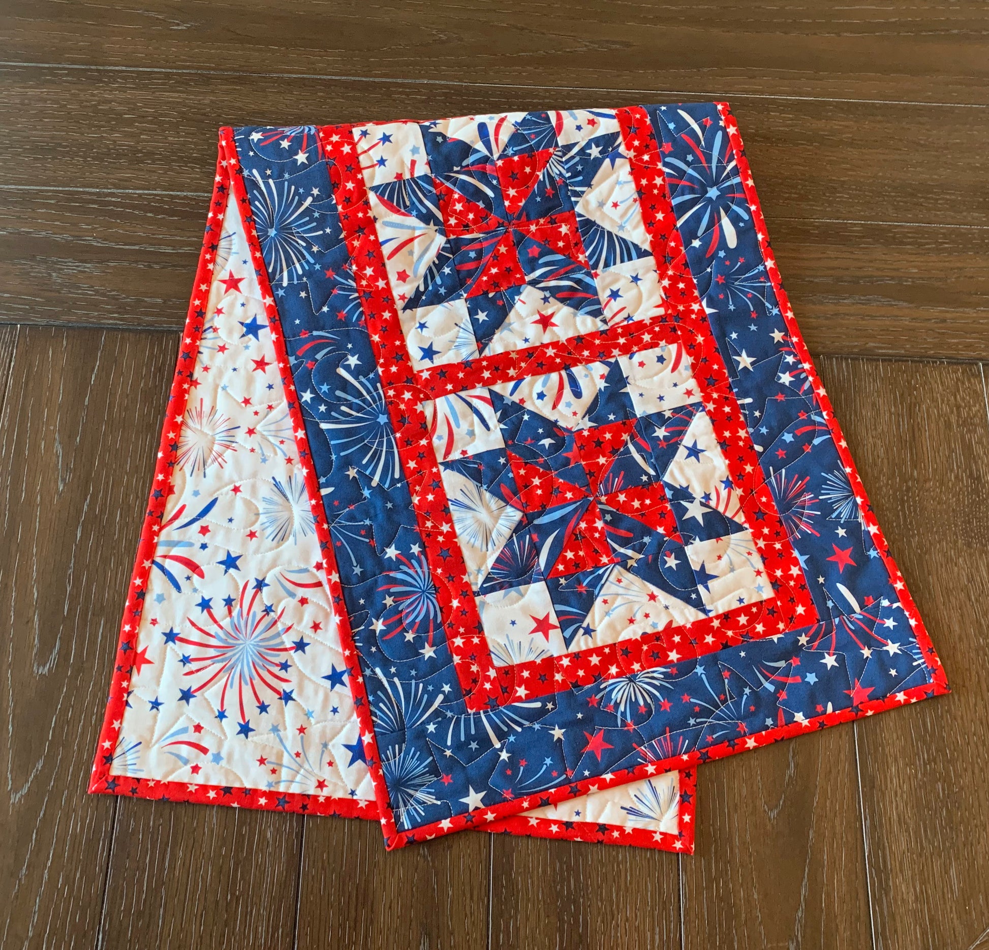 Red white and blue patriotic table runner pattern with four center star blocks that have a pinwheel in the center. The runner has red star sashing between the blocks and a blue fireworks border. Runner is shown folded on a table.