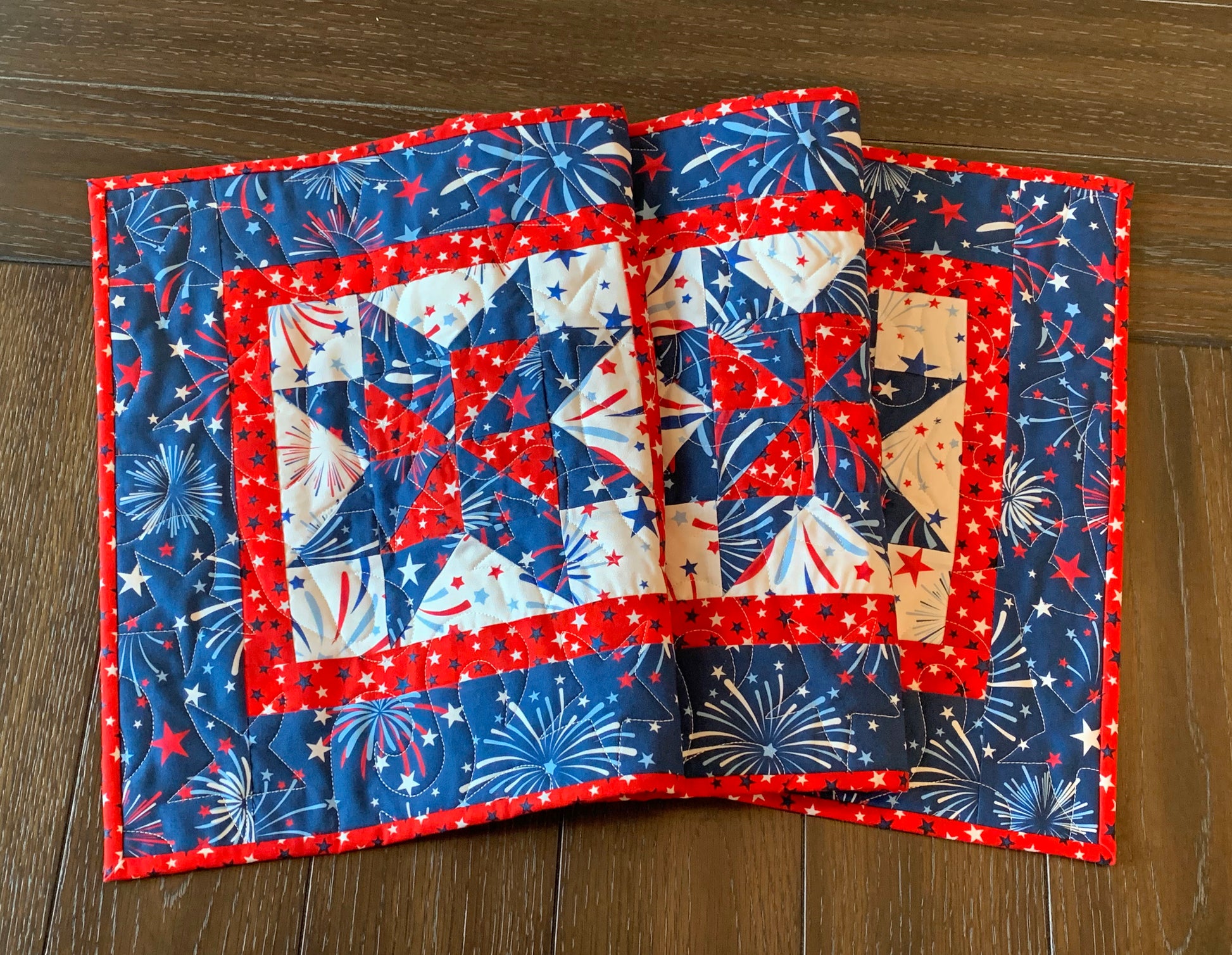 Red white and blue patriotic table runner with four center star blocks that have a pinwheel in the center. The runner has red star sashing between the blocks and a blue fireworks border. Runner is shown displayed on a table.