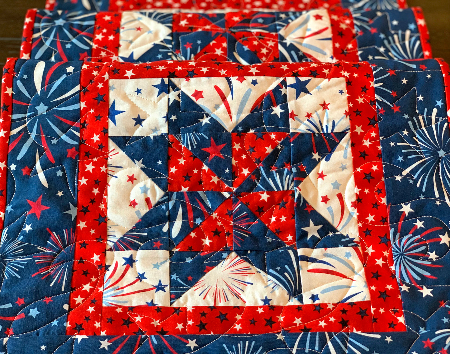 Close up of Red white and blue patriotic table runner pattern with four center star blocks that have a pinwheel in the center. The runner has red star sashing between the blocks and a blue fireworks border. 