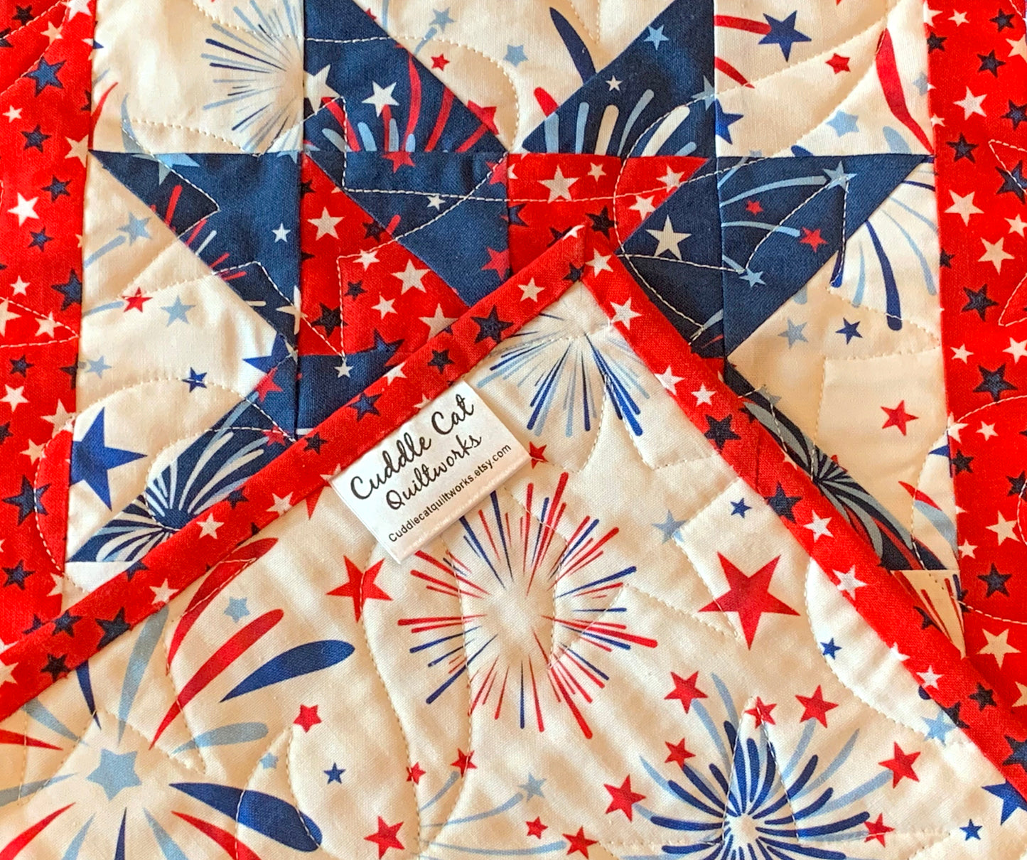 Close up of corner label on Red white and blue patriotic table runner with four center star blocks that have a pinwheel in the center. The runner has red star sashing between the blocks and a blue fireworks border. 