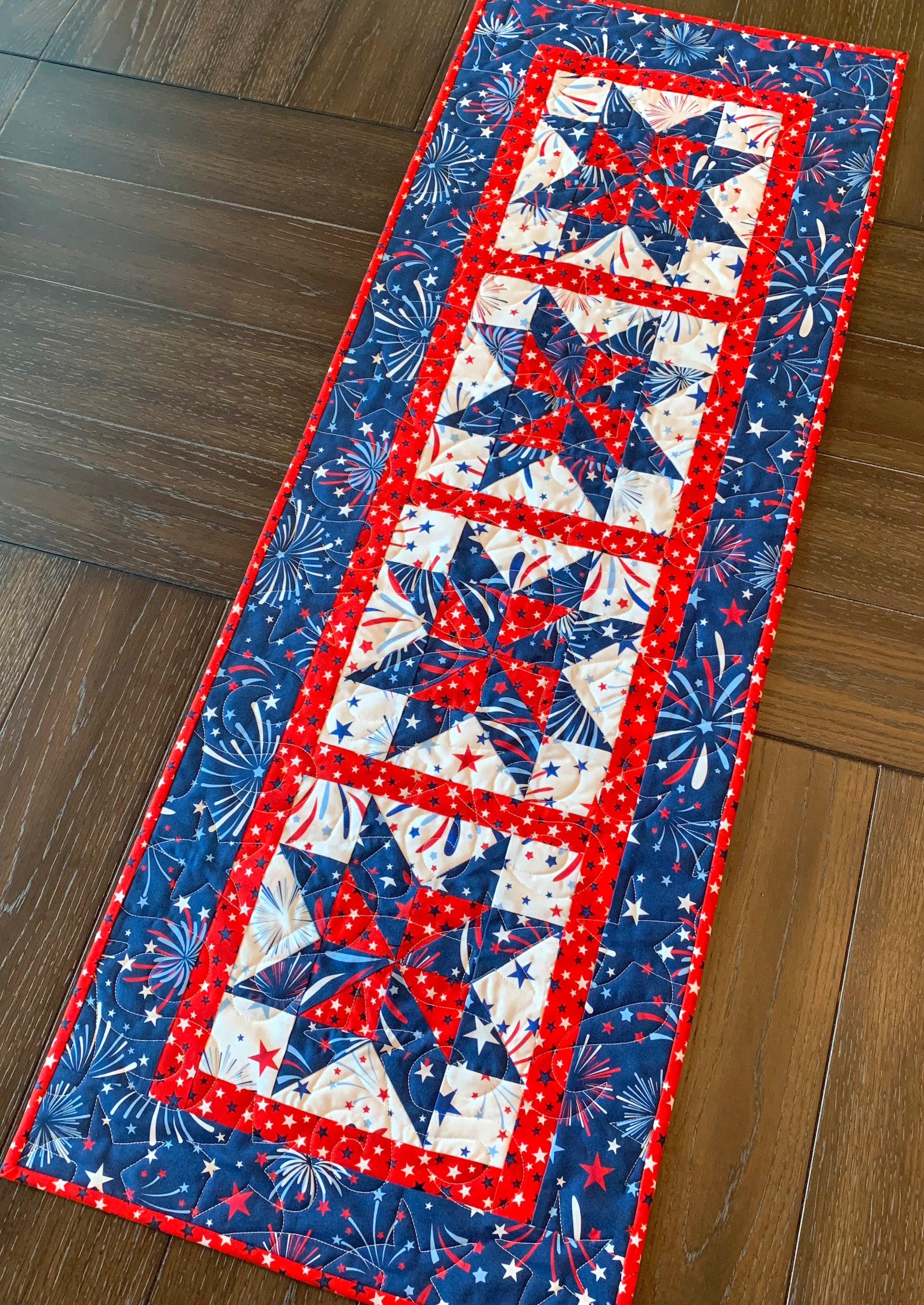 Red white and blue patriotic table runner with four center star blocks that have a pinwheel in the center. The runner has red star sashing between the blocks and a blue fireworks border. Runner is shown displayed on a table.