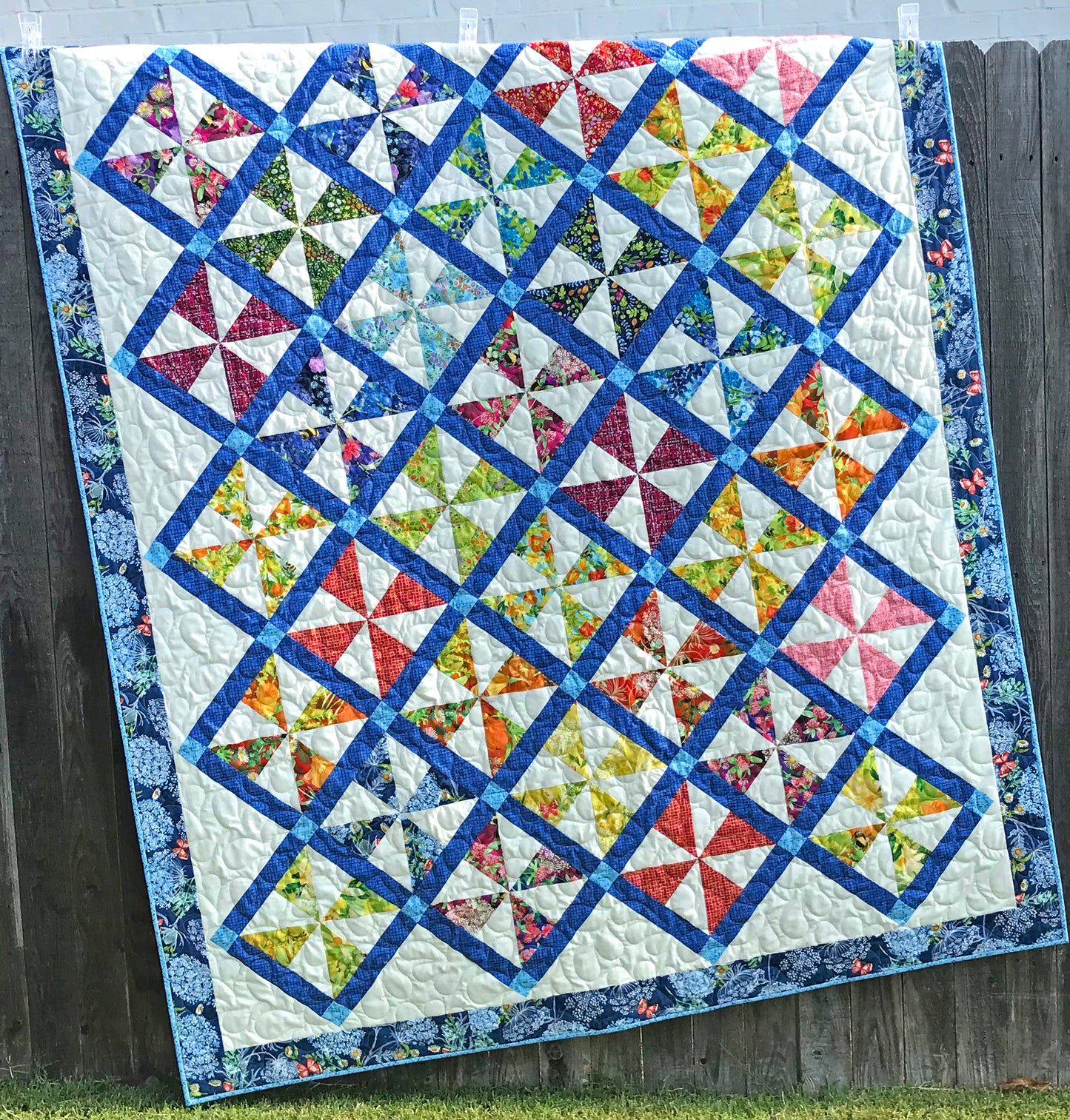 Pinwheel Parade quilt pattern featuring pinwheel blocks set on-point with sashing between the blocks and a floral border. Quilt is shown hanging on a fence.