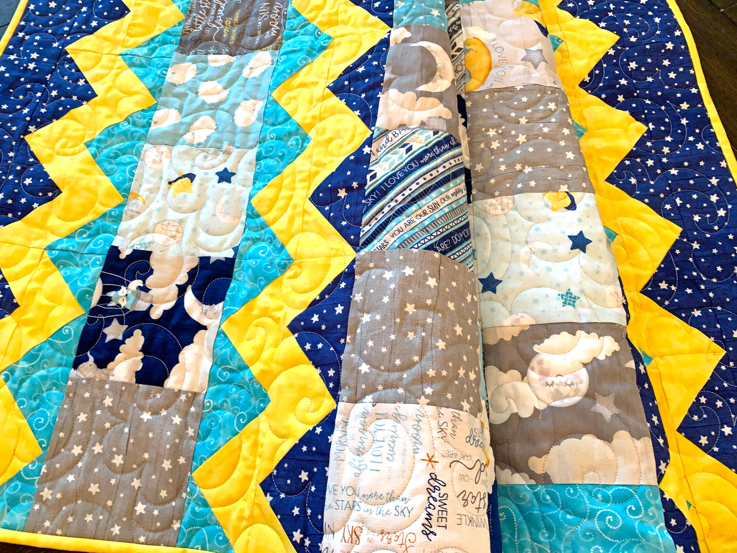 Close up of quilting on colorful charm square baby quilt pattern with yellow top and bottom zig-zag rows that look like rick-rack with rows of patchwork squares in between. Quilt is trimmed in blue and yellow and yellow.