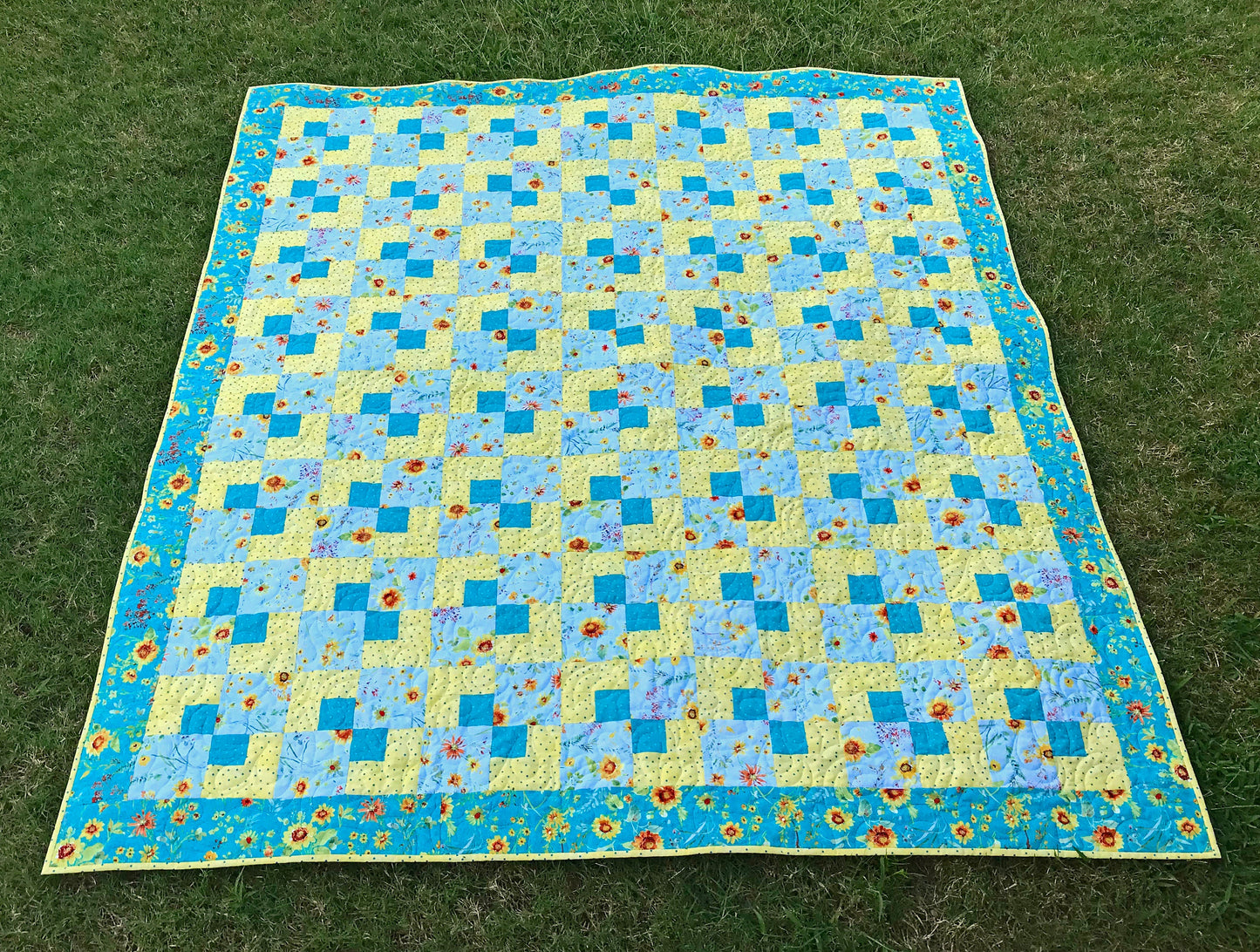 Cornerstones quilt pattern sample quilt. Four patch quilt in yellow and teal floral fabrics displayed laying on a lawn.