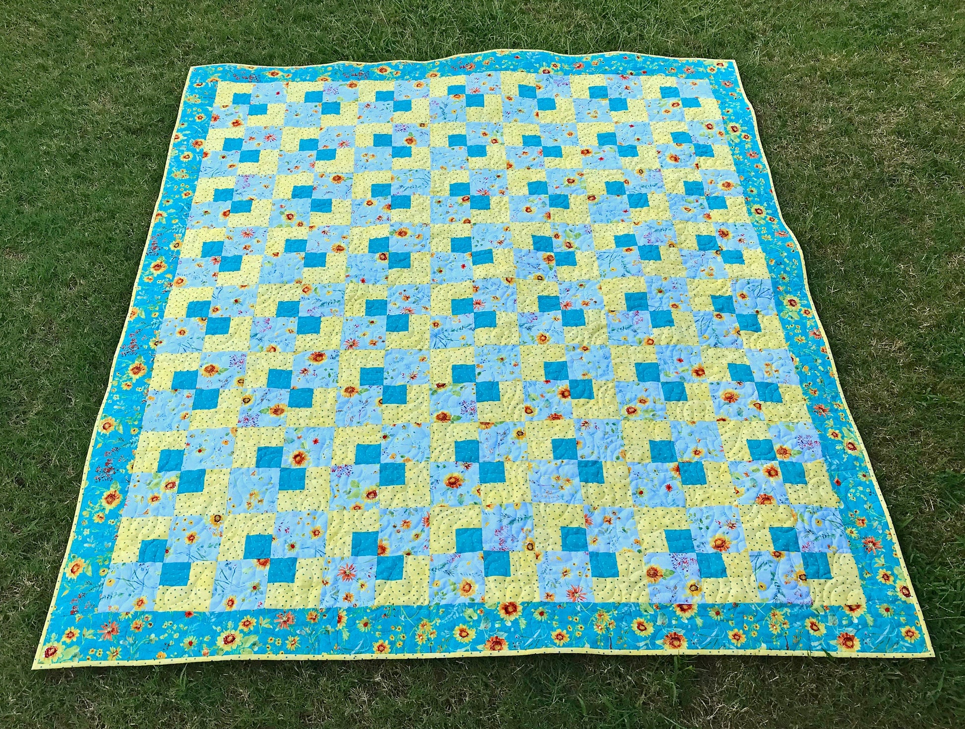 Cornerstones quilt pattern sample quilt. Four patch quilt in yellow and teal floral fabrics displayed laying on a lawn.