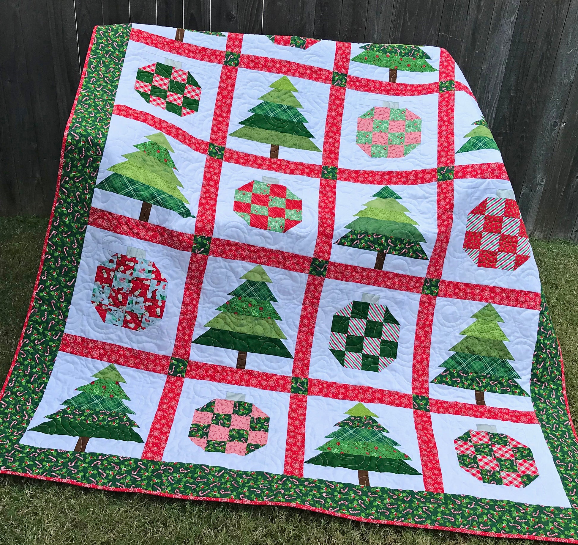 Christmastime quilt pattern with Christmas trees and ornament blocks surrounded by red sashing and a green holly border. Quilt is shown laying on a bench