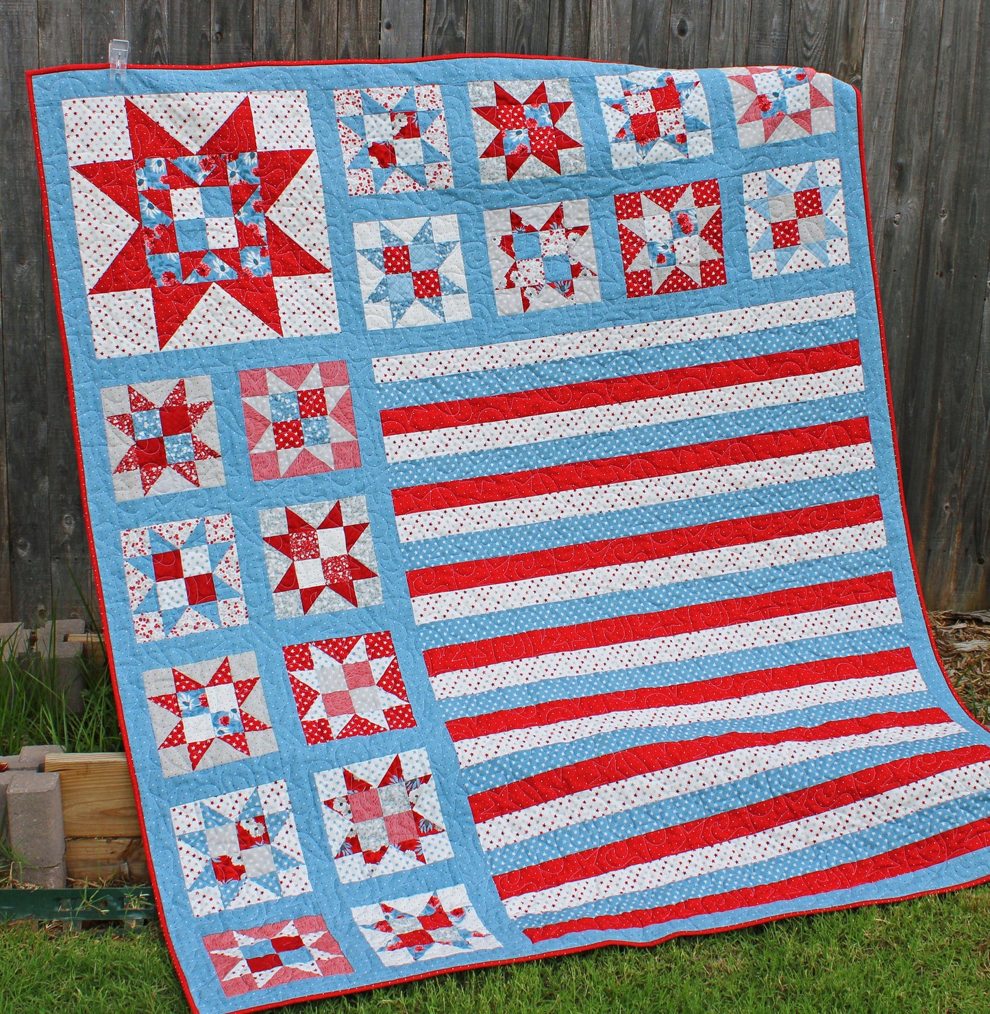 One Nation patriotic quilt pattern sample quilt with patchwork stars in three sizes and red white and blue stripes and blue sashing. Quilt is shown hanging on a fence.