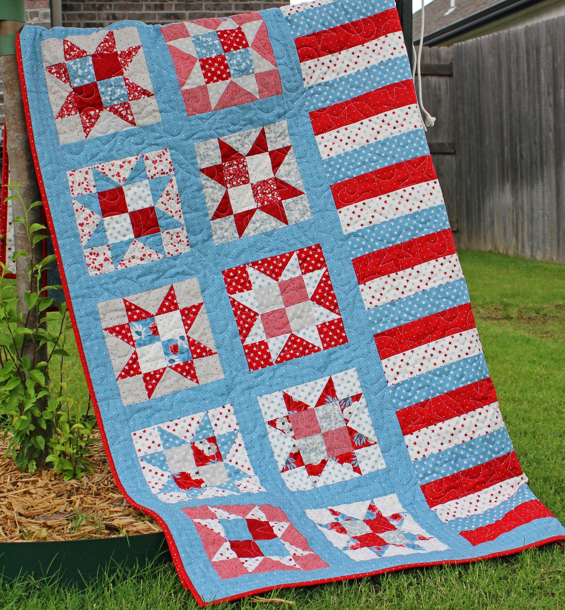 One Nation patriotic quilt pattern sample quilt with patchwork stars in three sizes and red white and blue stripes and blue sashing. Quilt is shown folded over and hanging on a fence with the stars and stripes showing.