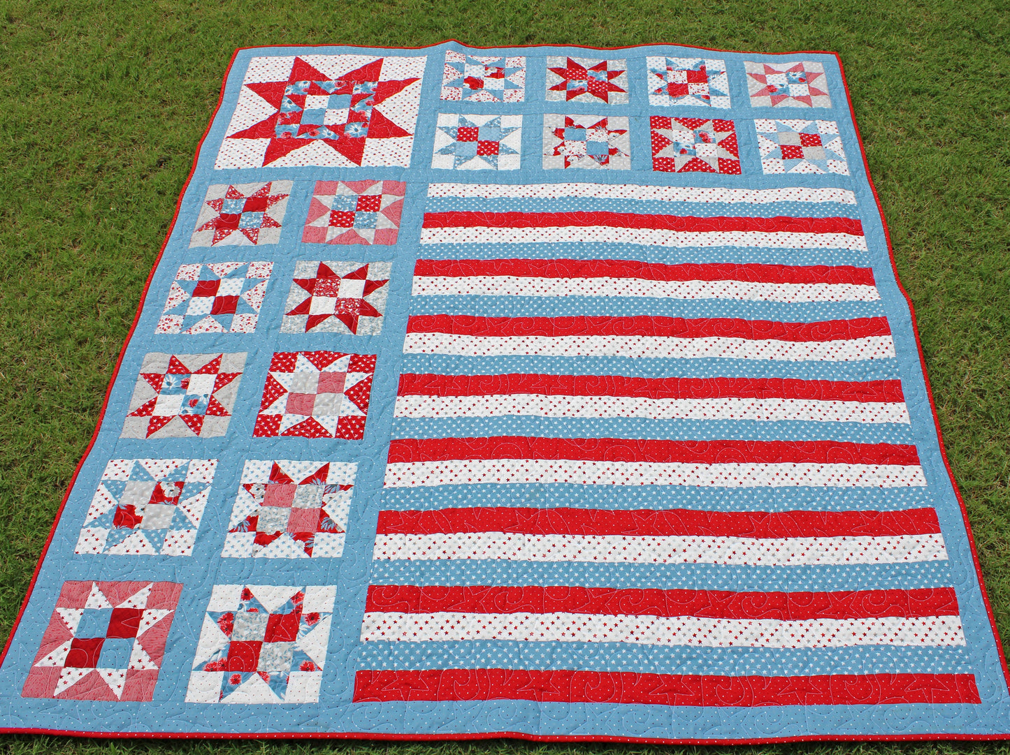 One Nation patriotic quilt pattern sample quilt with patchwork stars in three sizes and red white and blue stripes and blue sashing. Quilt is shown laying on the grass.