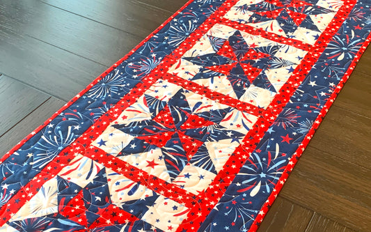 Red white and blue patriotic table runner with four center star blocks that have a pinwheel in the center. The runner has red star sashing between the blocks and a blue fireworks border. Runner is shown displayed on a table.