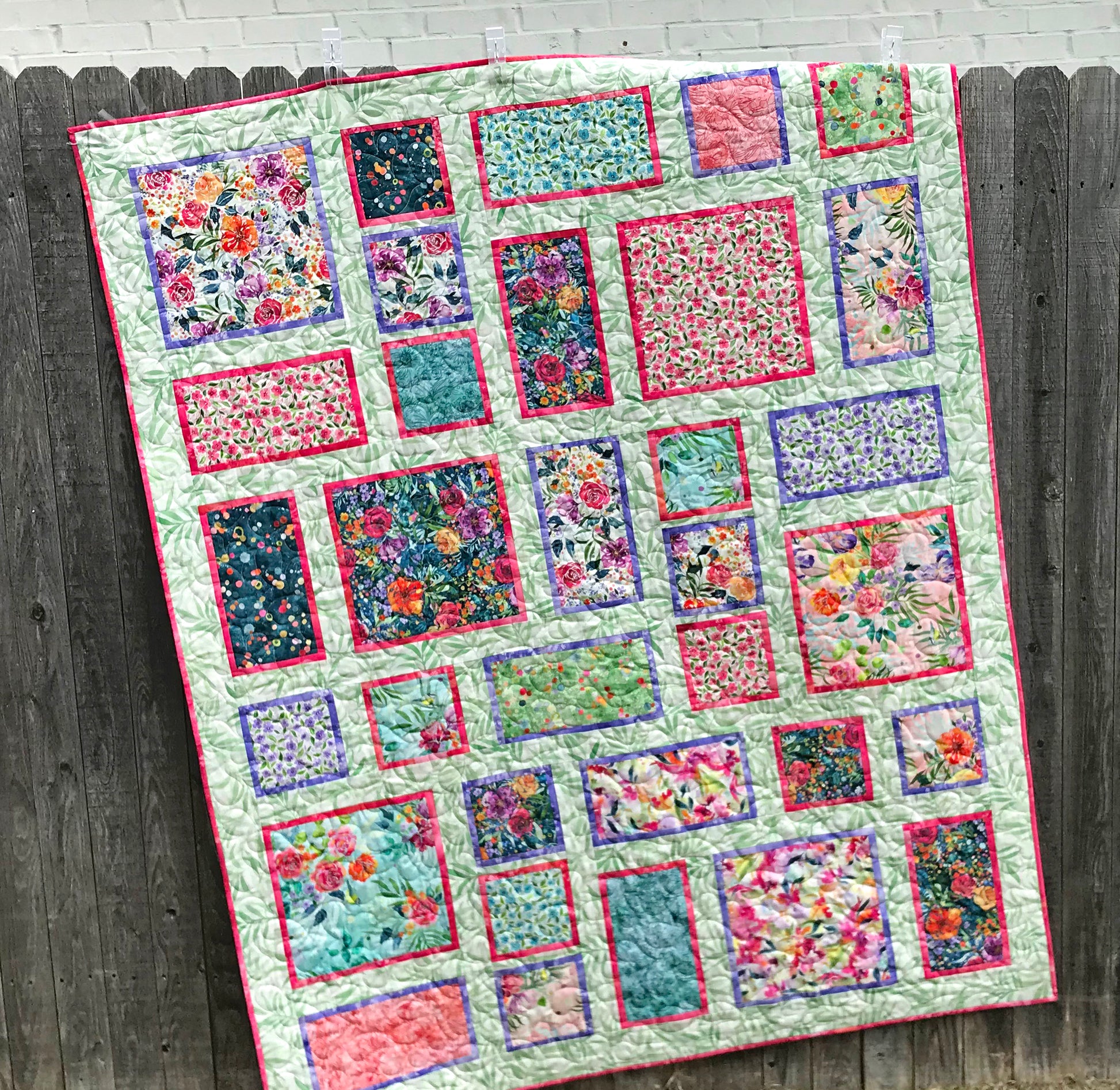 Handmade quilt featuring watercolor floral squares and rectangles framed with pink and purple fabric on a light green background. Quilt is shown hanging on a fence.