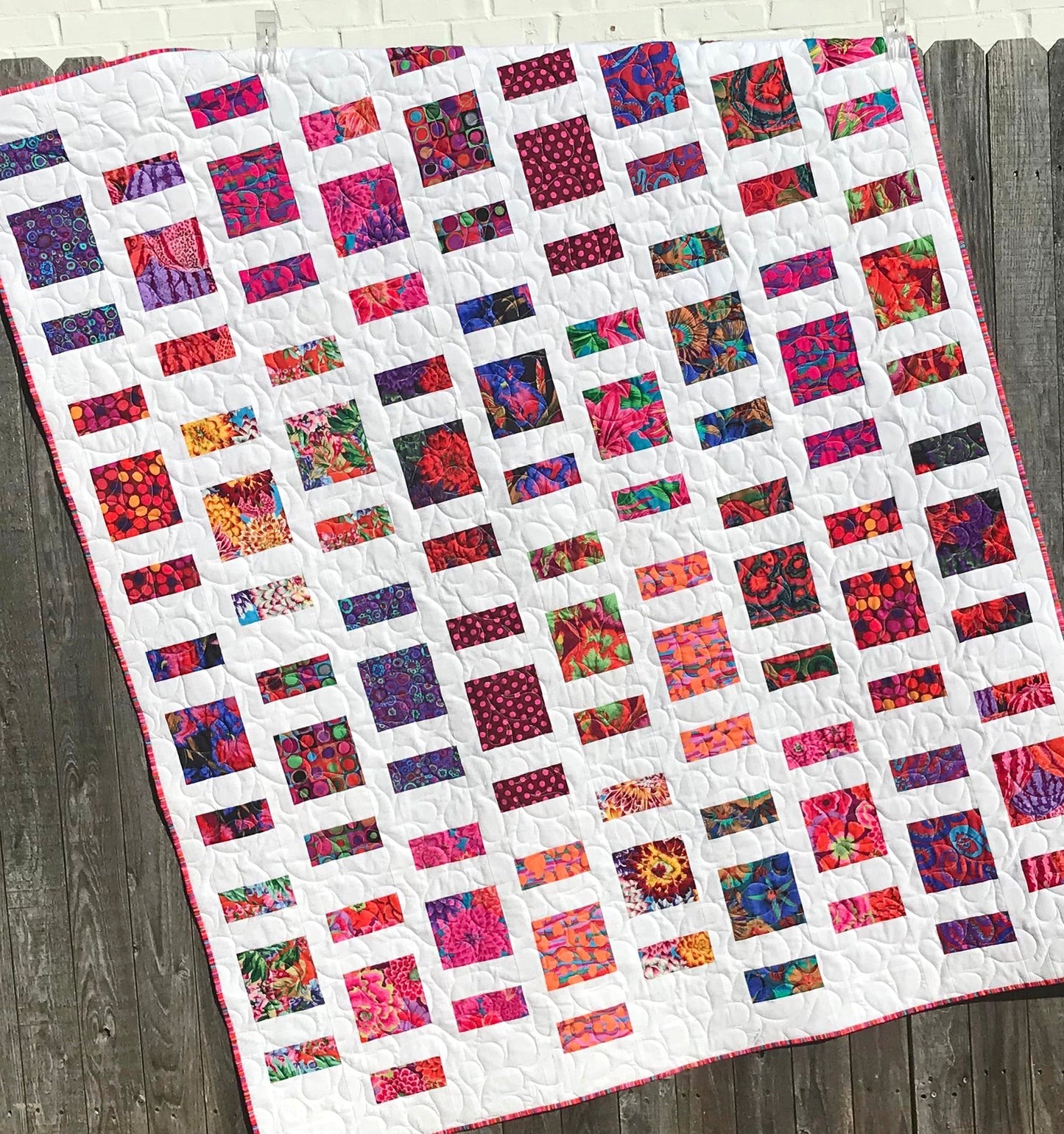 Quilt Pattern for charm squares that has nine columns of squares and rectangles with sashing between the columns. Quilt is displayed on a fence with colorful Kaffe Fassett fabrics.