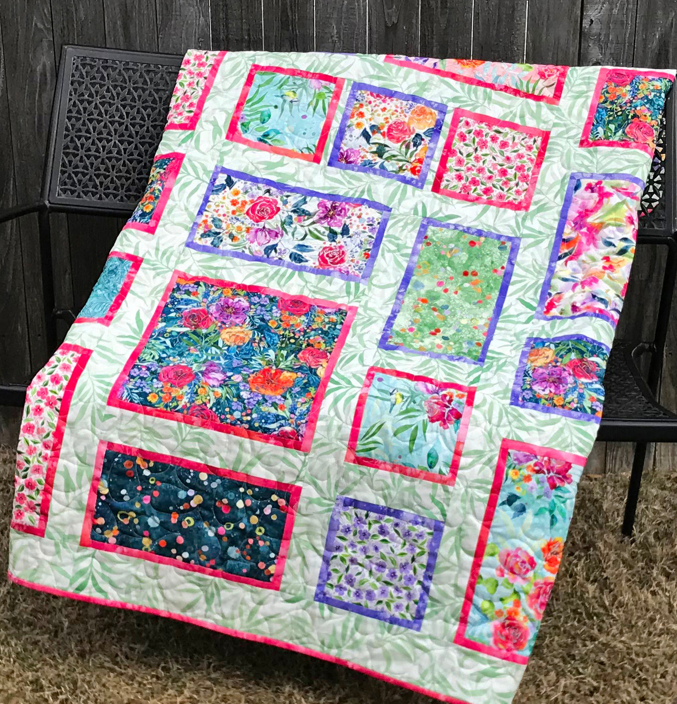 Handmade quilt featuring watercolor floral squares and rectangles framed with pink and purple fabric on a light green background. Quilt is shown displayed on a bench.