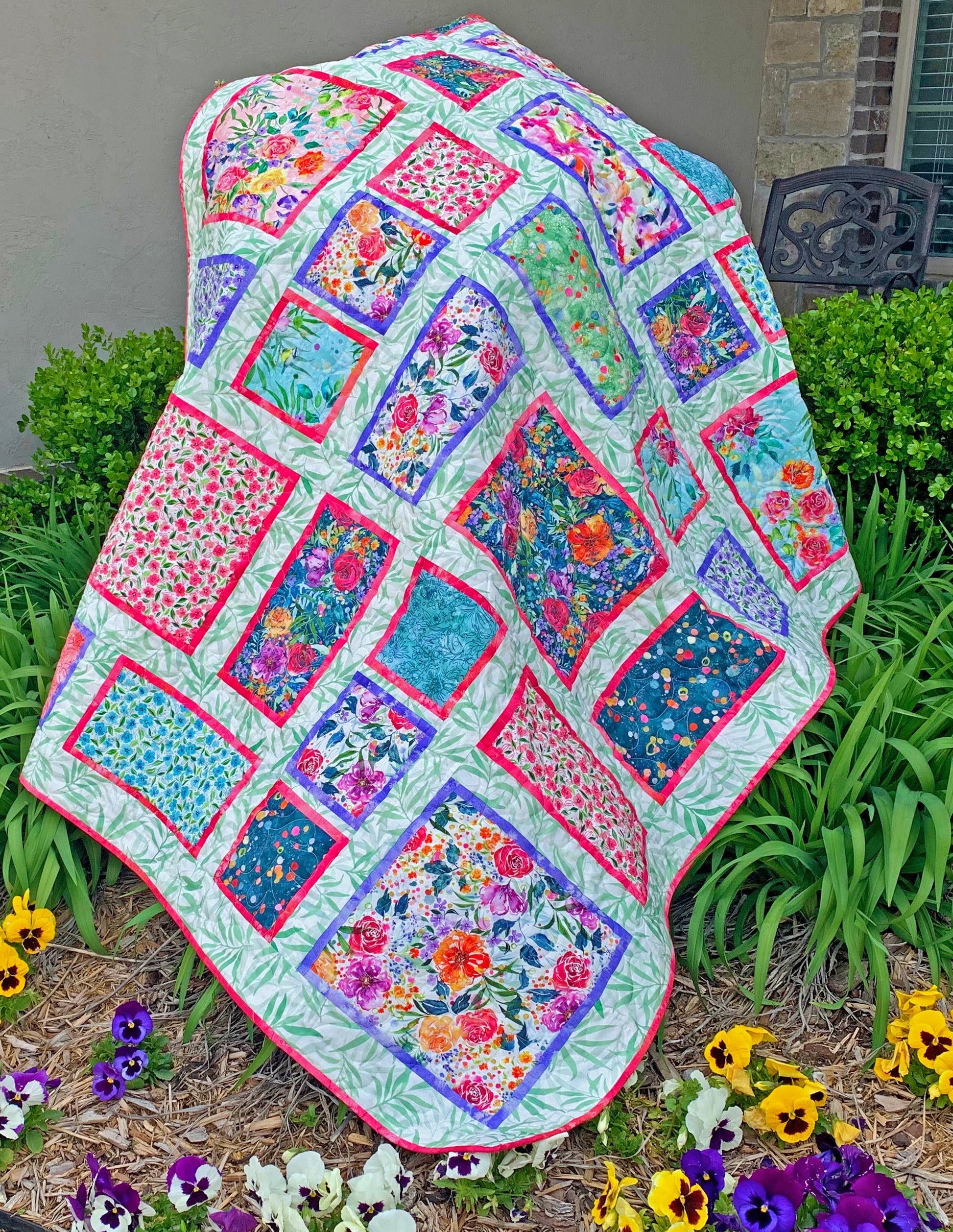 Fancy Frames Quilt pattern of watercolor floral squares and rectangles framed with pink and purple fabric on a light green background. Close up of quilt is displayed.