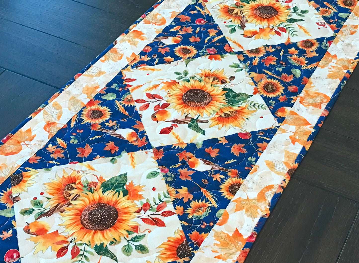 Sunflower themed fall table runner with a center diamond pattern of sunflowers surrounded by gold leaves and dark blue accents displayed on a table