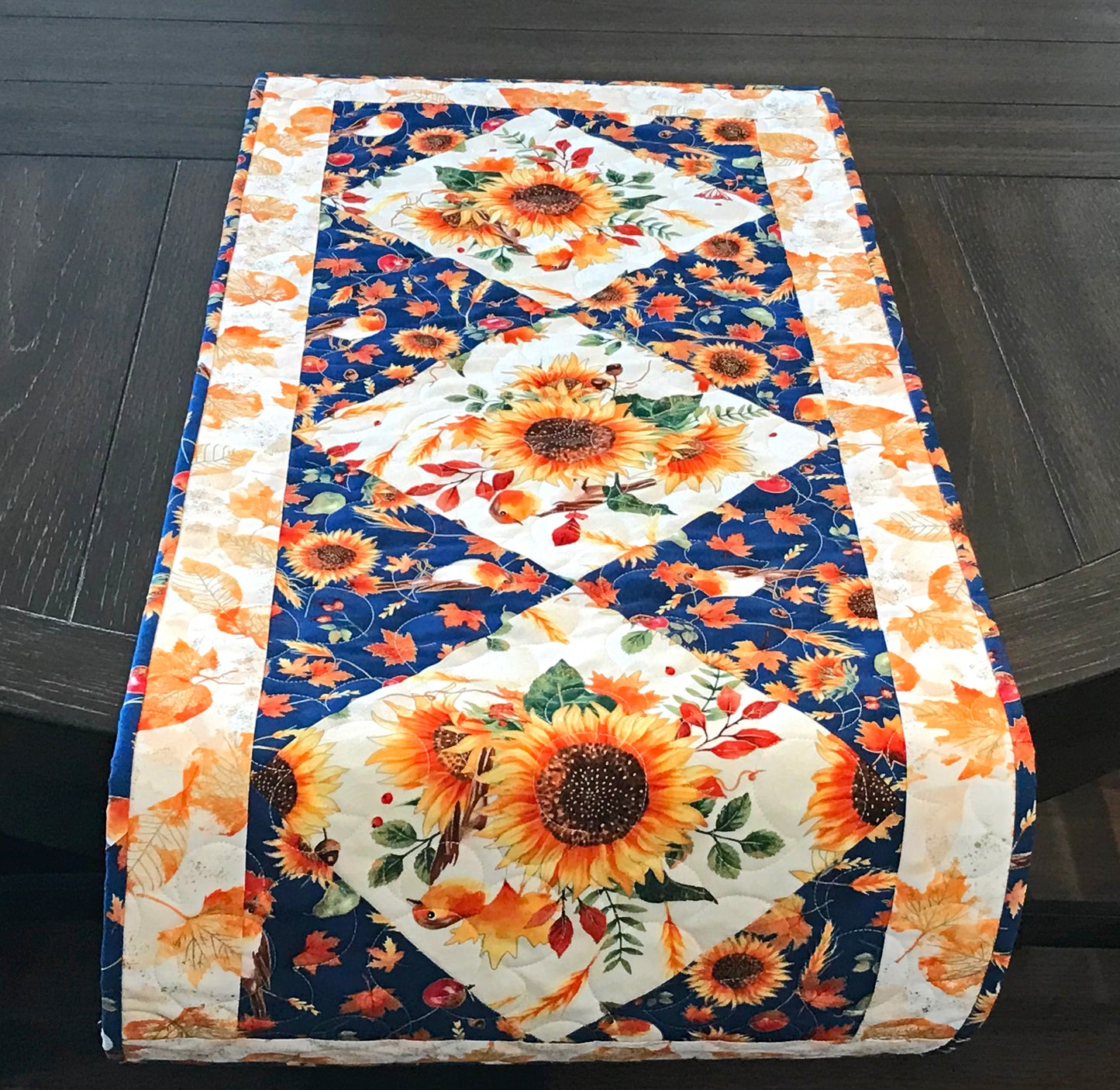 Sunflower themed fall table runner with a center diamond pattern of sunflowers surrounded by gold leaves and dark blue accents displayed on a table