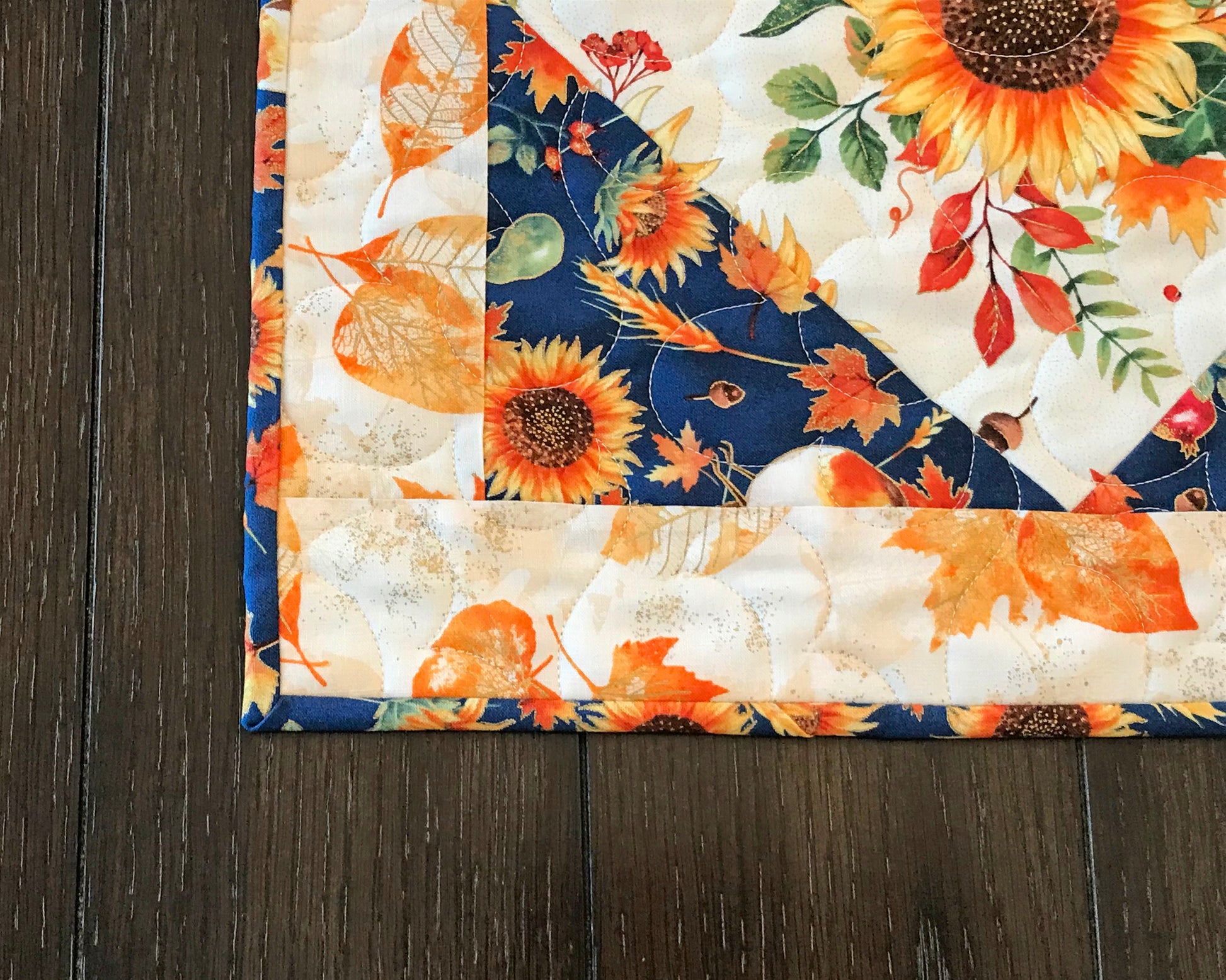 corner close up of Sunflower themed fall table runner with a center diamond pattern of sunflowers surrounded by gold leaves and dark blue accents displayed on a table