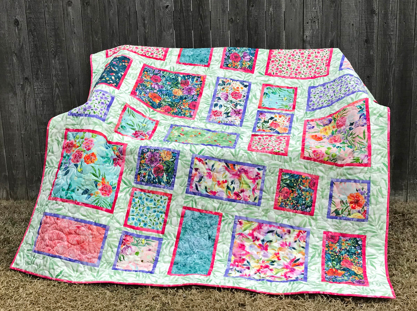 Handmade quilt featuring watercolor floral squares and rectangles framed with pink and purple fabric on a light green background. Quilt is shown displayed on a bench.