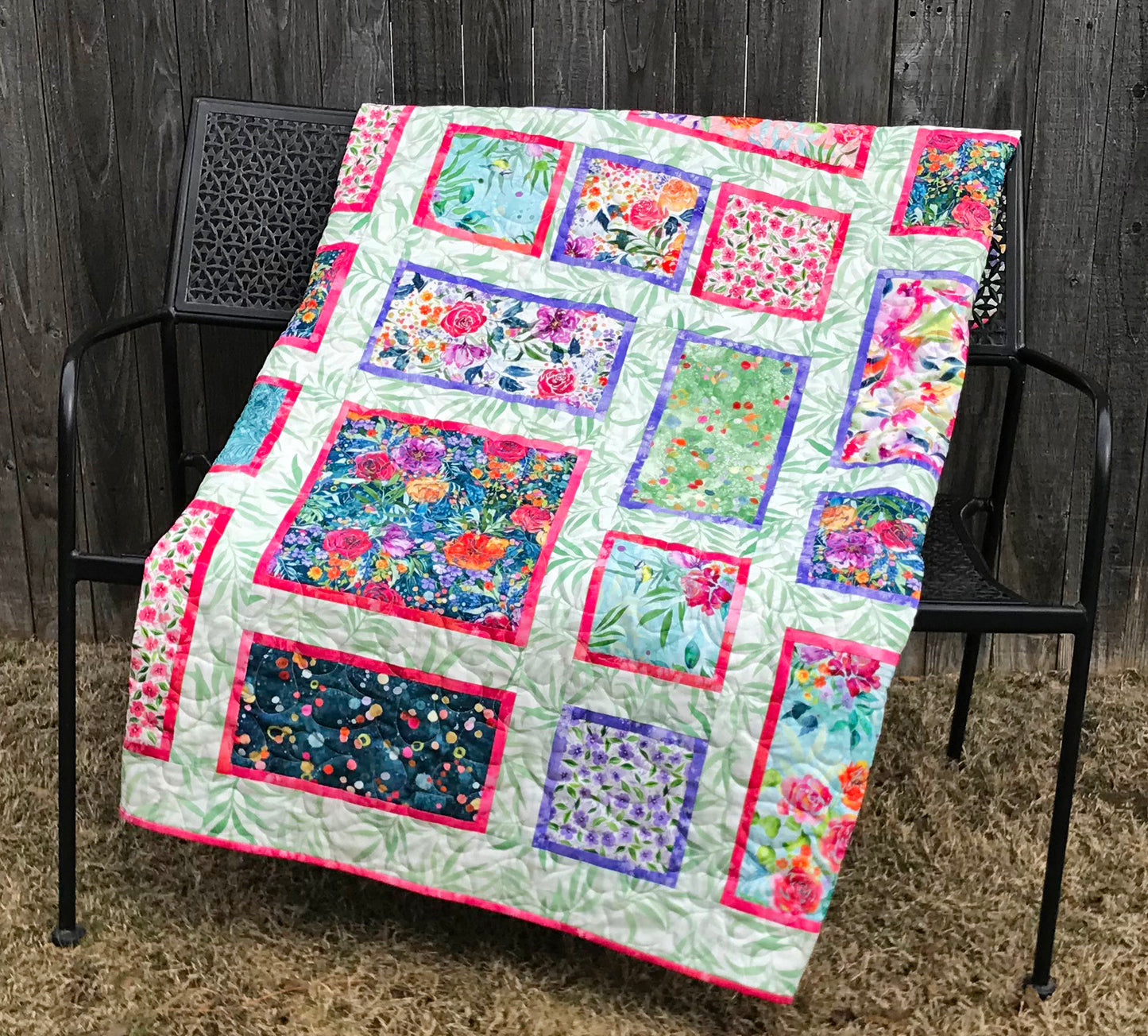 Handmade quilt featuring watercolor floral squares and rectangles framed with pink and purple fabric on a light green background. Quilt is shown displayed on a bench.