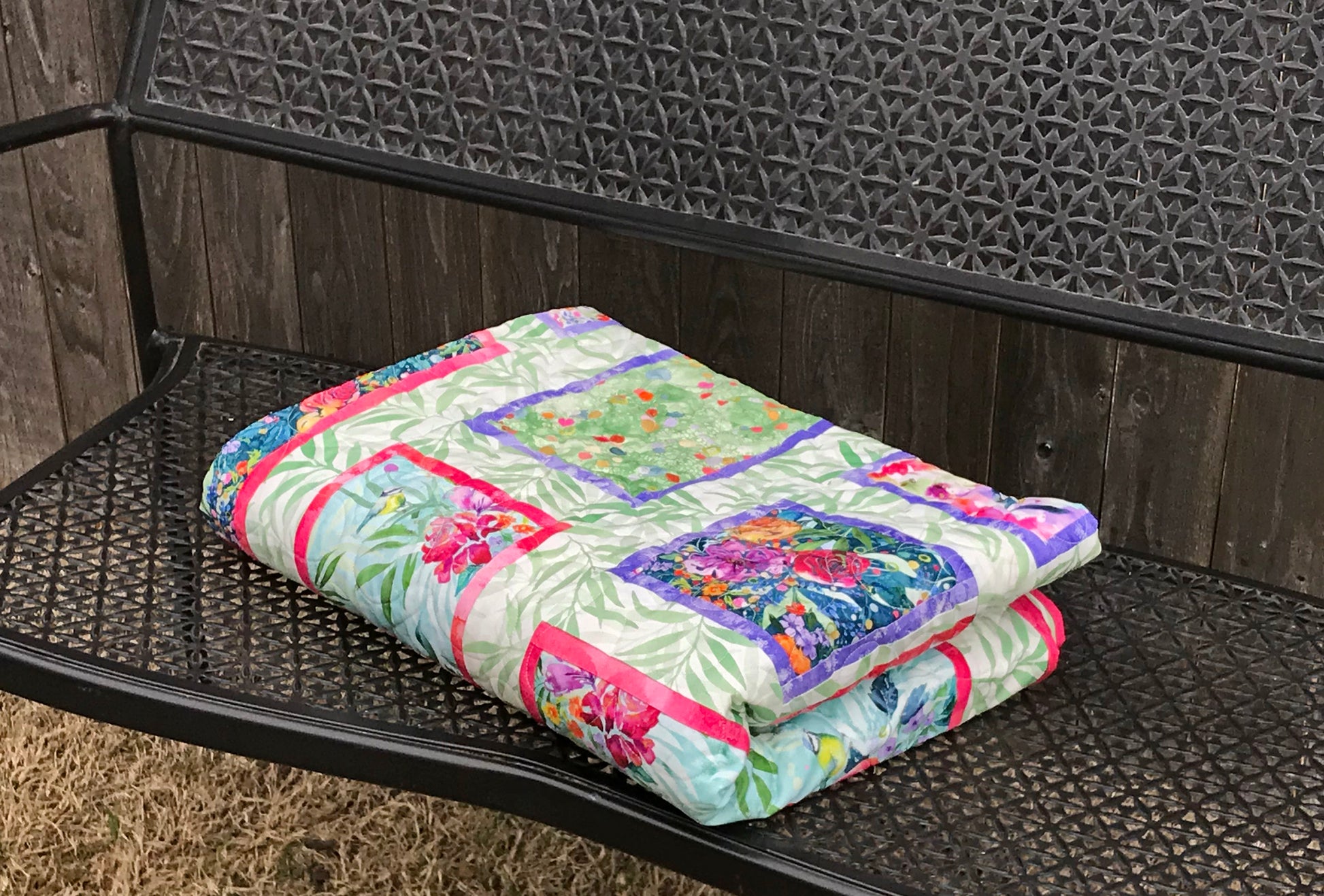 Handmade quilt featuring watercolor floral squares and rectangles framed with pink and purple fabric on a light green background. Quilt is shown folded on a bench.