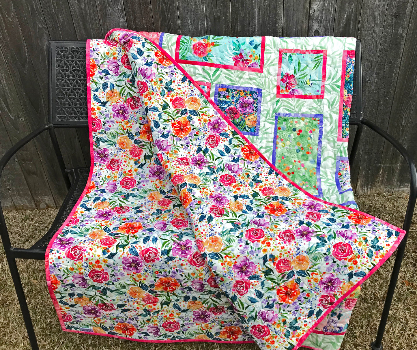 Handmade quilt featuring watercolor floral squares and rectangles framed with pink and purple fabric on a light green background. Quilt is shown folded on a bench. with the floral backing showing.