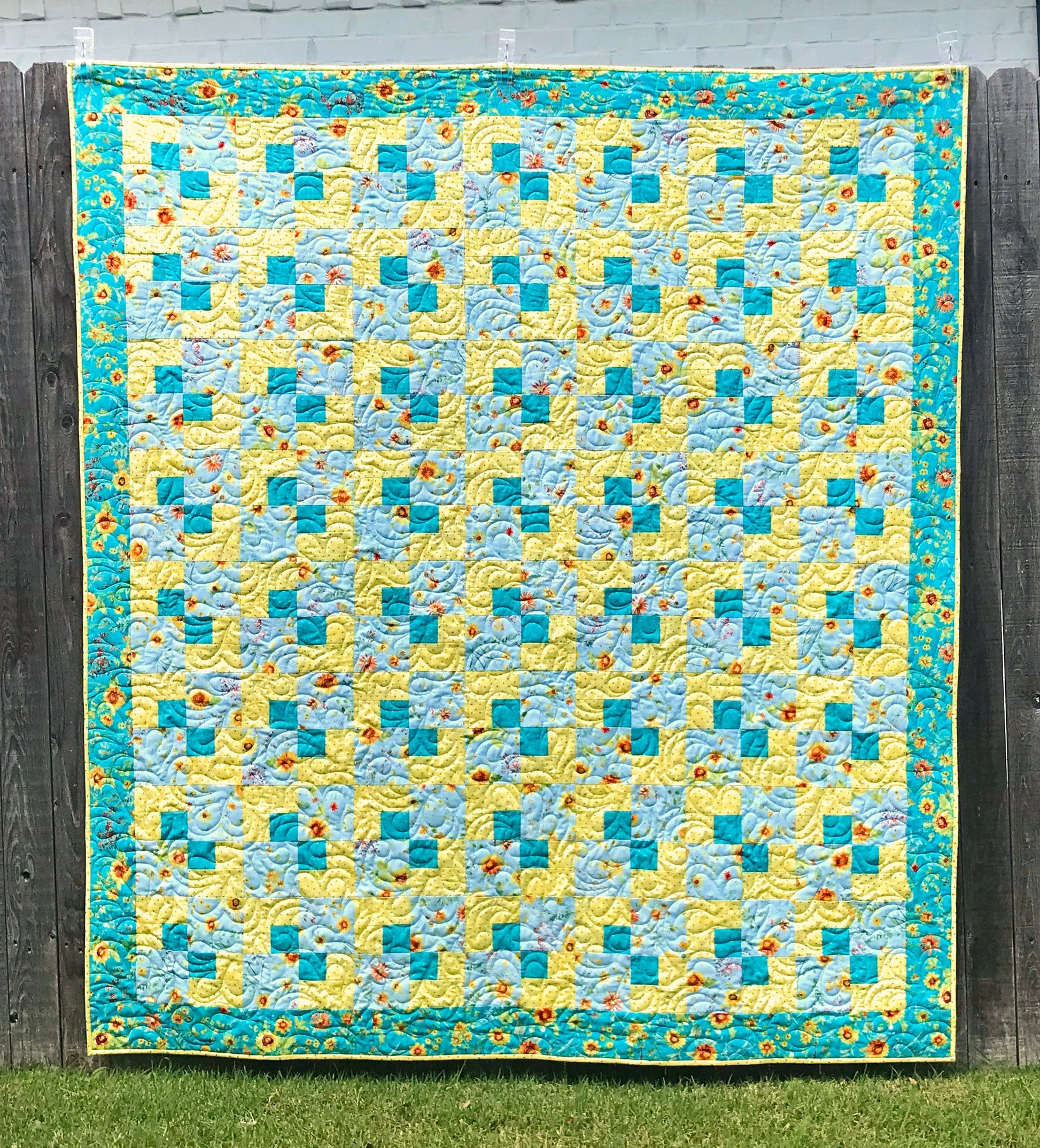 Cornerstones quilt pattern sample quilt. Four patch quilt in yellow and teal floral fabrics displayed on a fence.