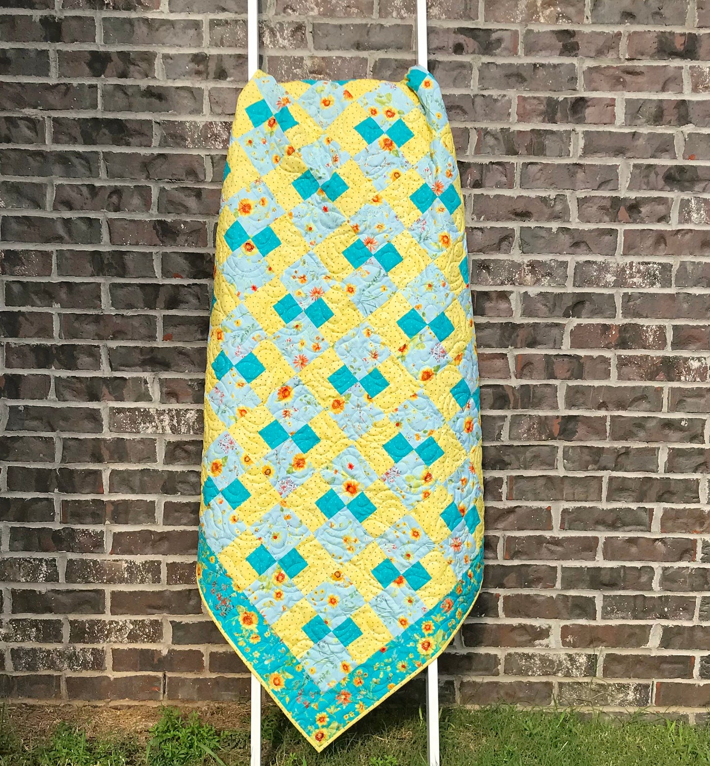 Cornerstones quilt pattern sample quilt. Four patch quilt in yellow and teal floral fabrics displayed on a ladder.