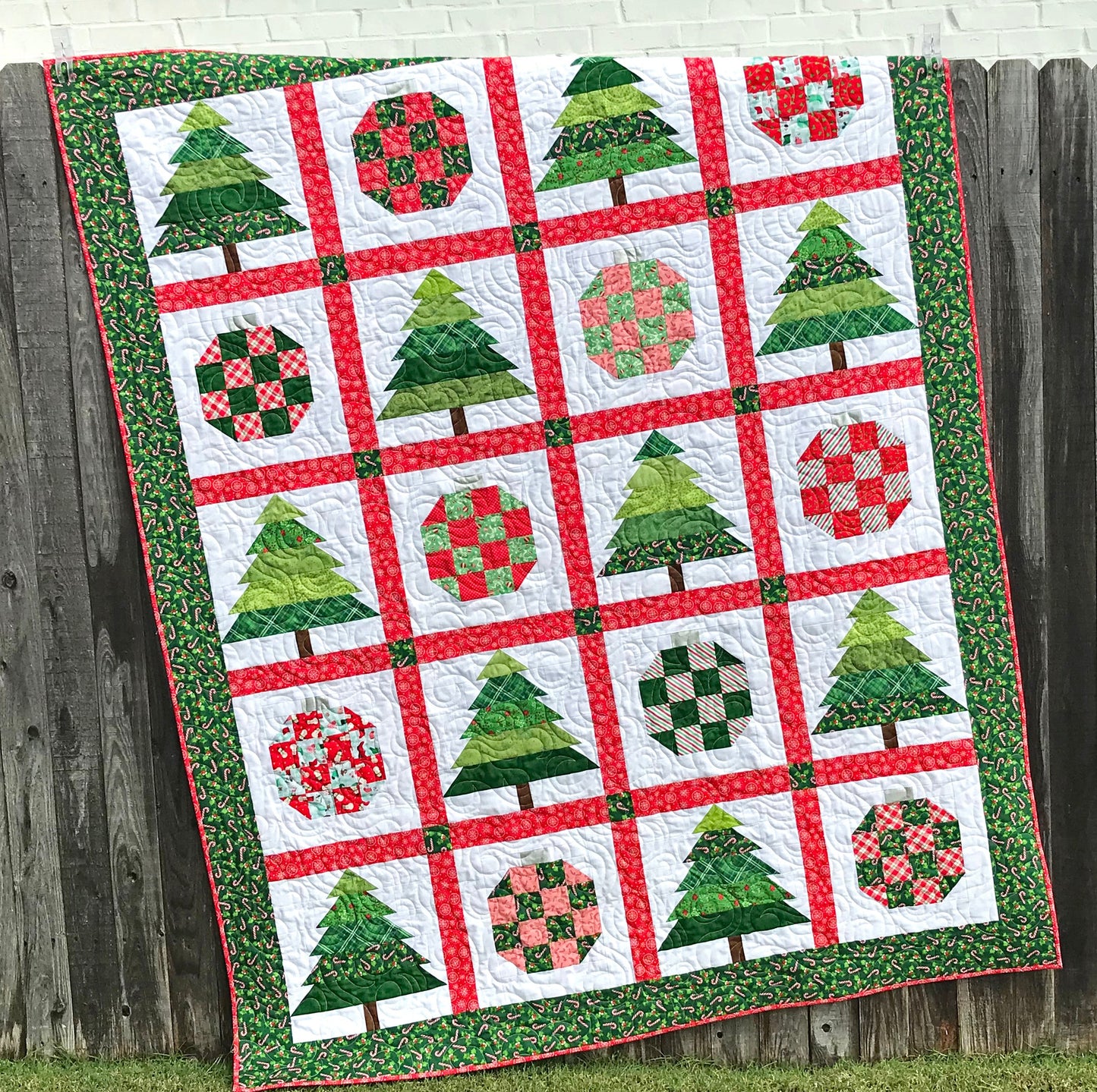 Christmastime quilt pattern with Christmas trees and ornament blocks surrounded by red sashing and a green holly border. Quilt is shown hanging on a fence.