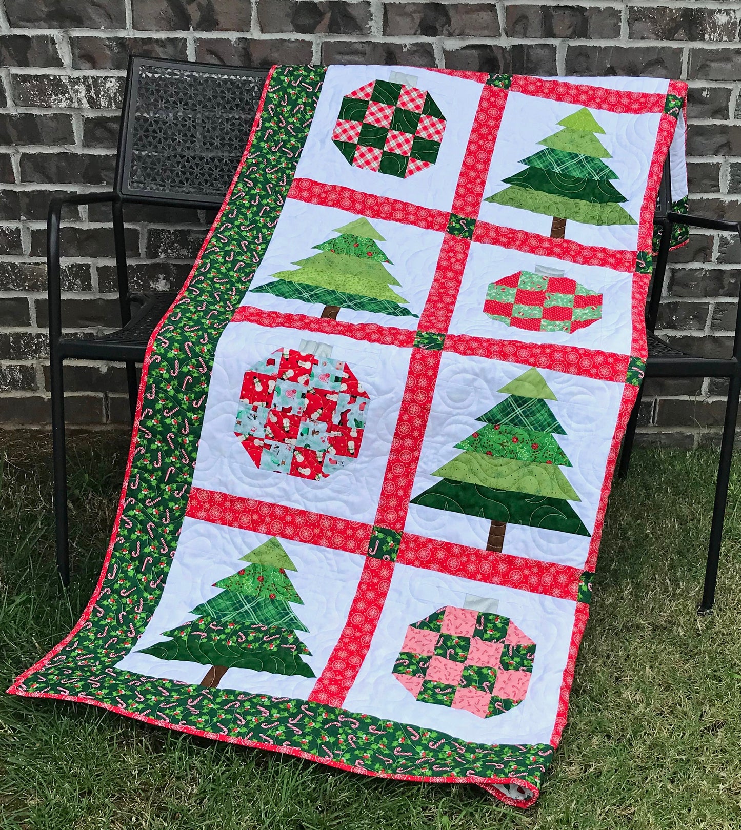 Christmastime quilt pattern with Christmas trees and ornament blocks surrounded by red sashing and a green holly border. Quilt is shown hanging on a fence.