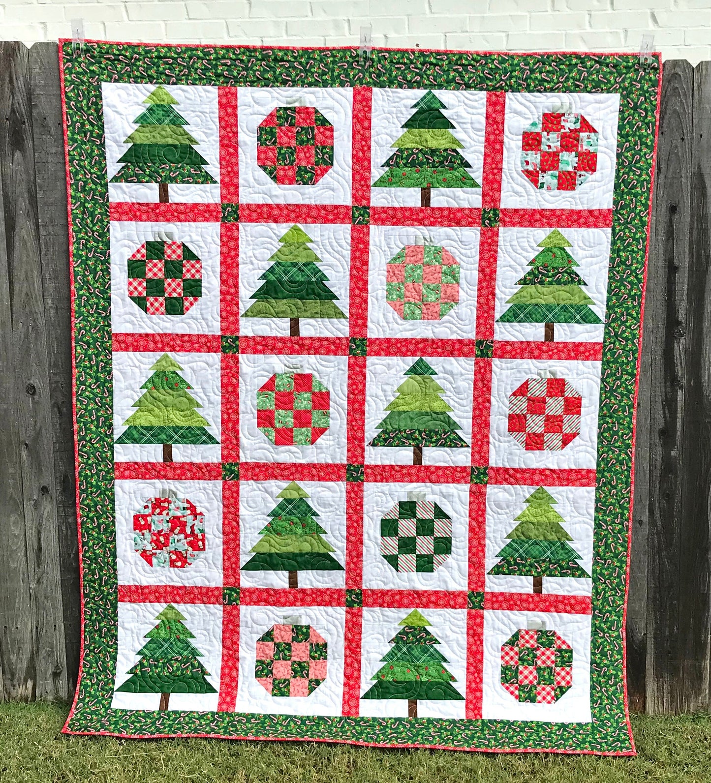 Christmastime quilt pattern with Christmas trees and ornament blocks surrounded by red sashing and a green holly border. Quilt is shown hanging on a fence.