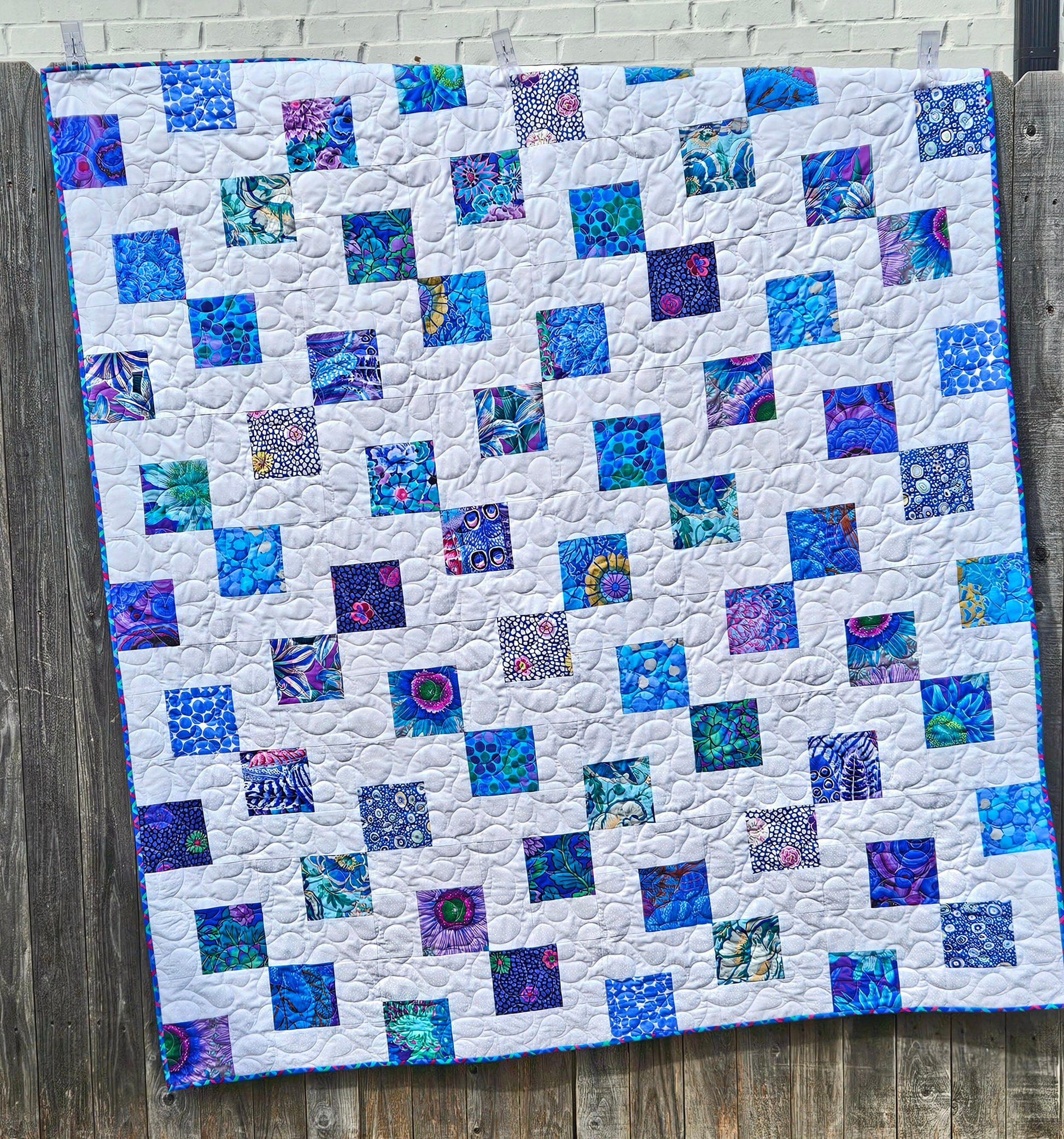 Double the Charm quilt pattern with sample quilt done in blue Kaffe Fassett charm squares on a white background. Quilt is shown hanging on a fence.